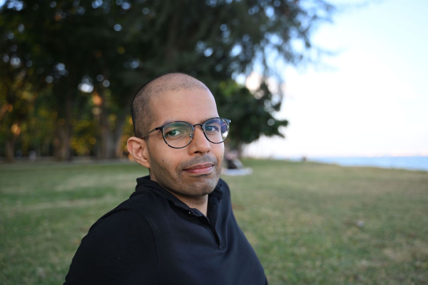 Bald man with glasses in black shirt poses outdoors near palm trees by the ocean.