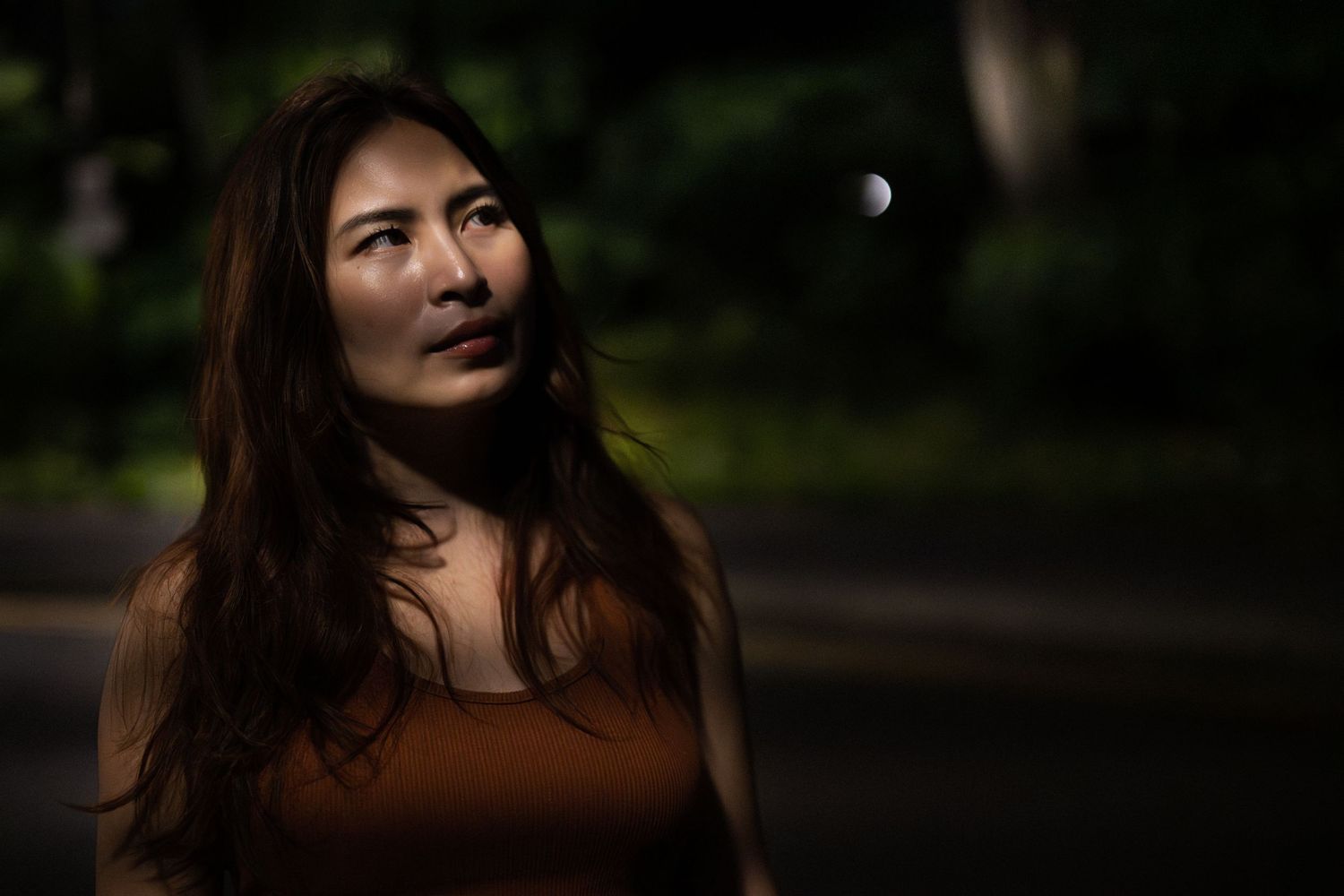 Young Asian woman in brown top gazes upward with moon visible in dark outdoor background.