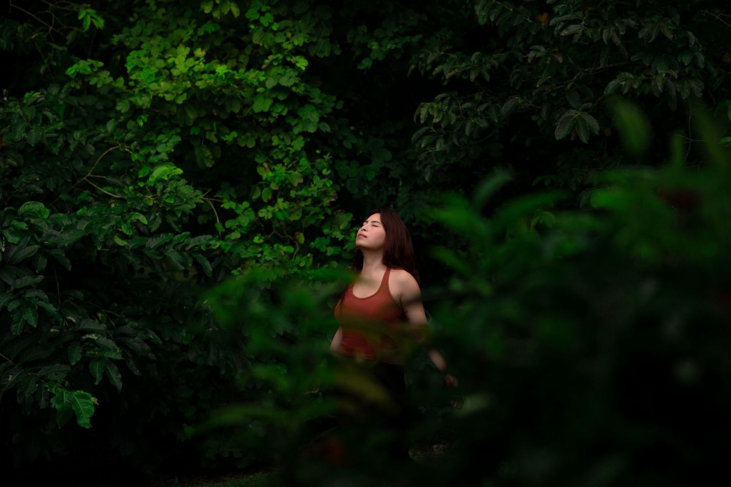 Woman in a red top standing among dense green tropical foliage with eyes closed.