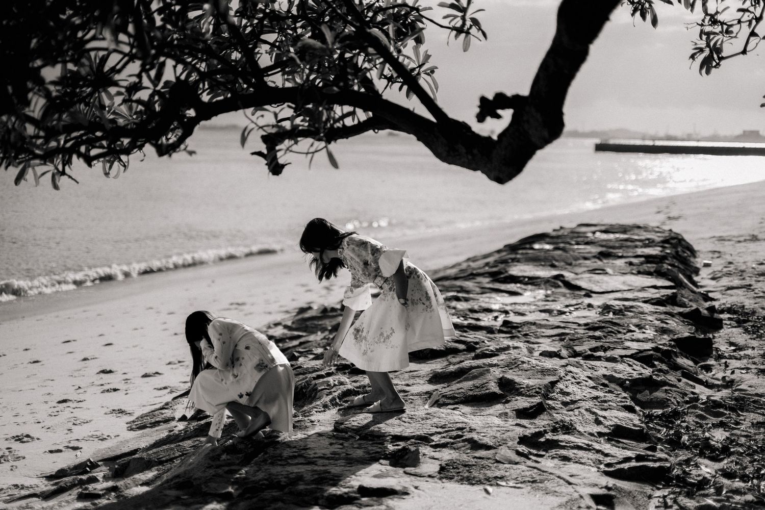 Black and white photo of two women in white dresses exploring rocks on a beach shoreline.