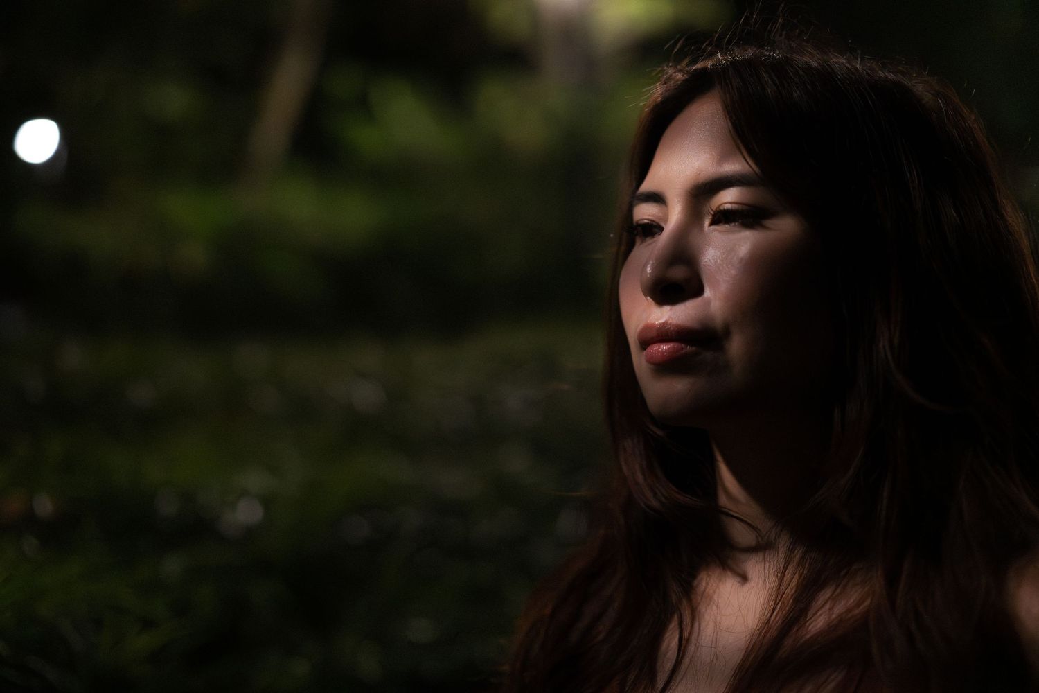 Close-up portrait of a woman with long hair in a dark outdoor garden setting at night.