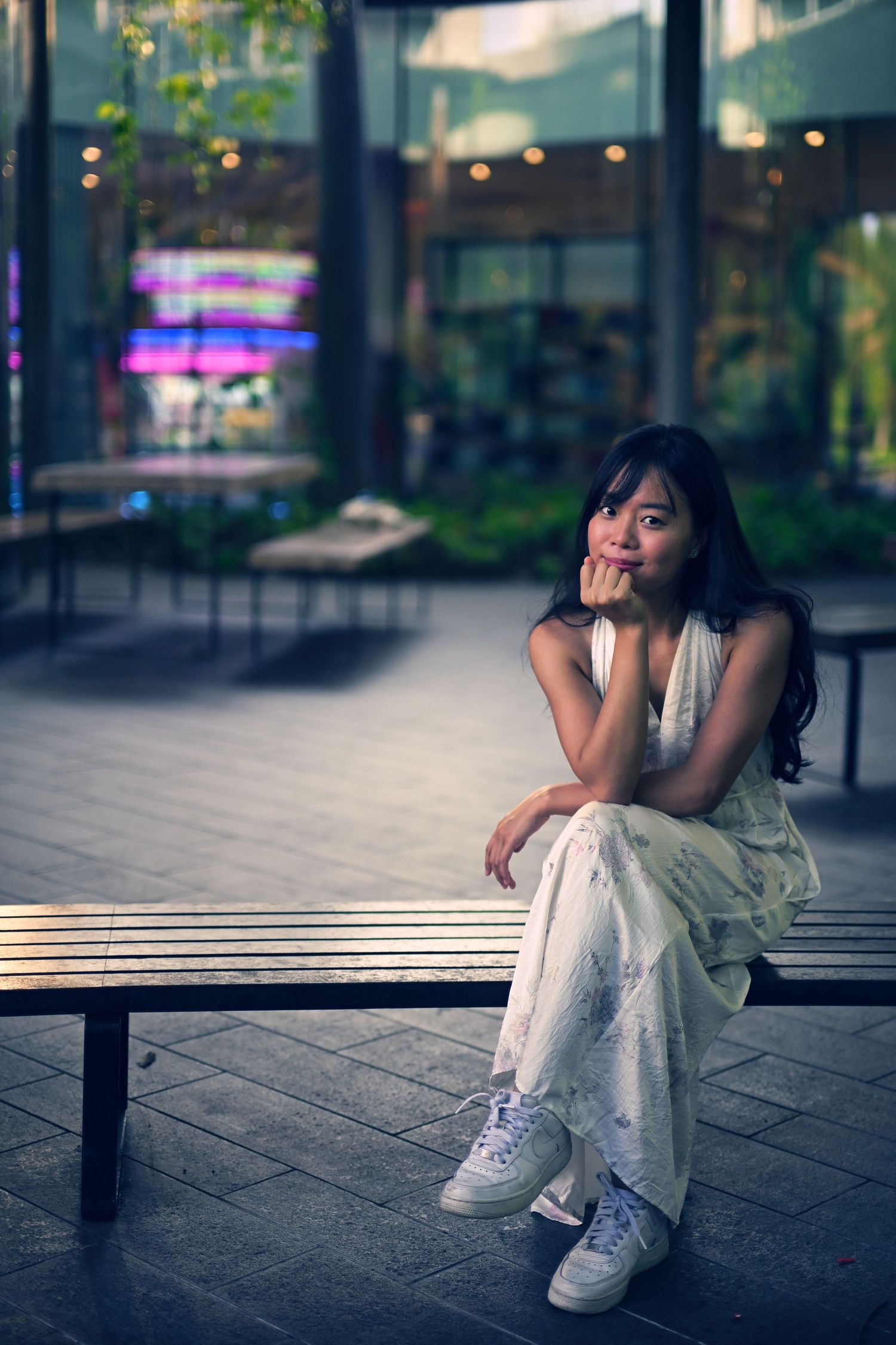 Young Asian woman in white floral dress sits pensively on a bench outside a modern building.