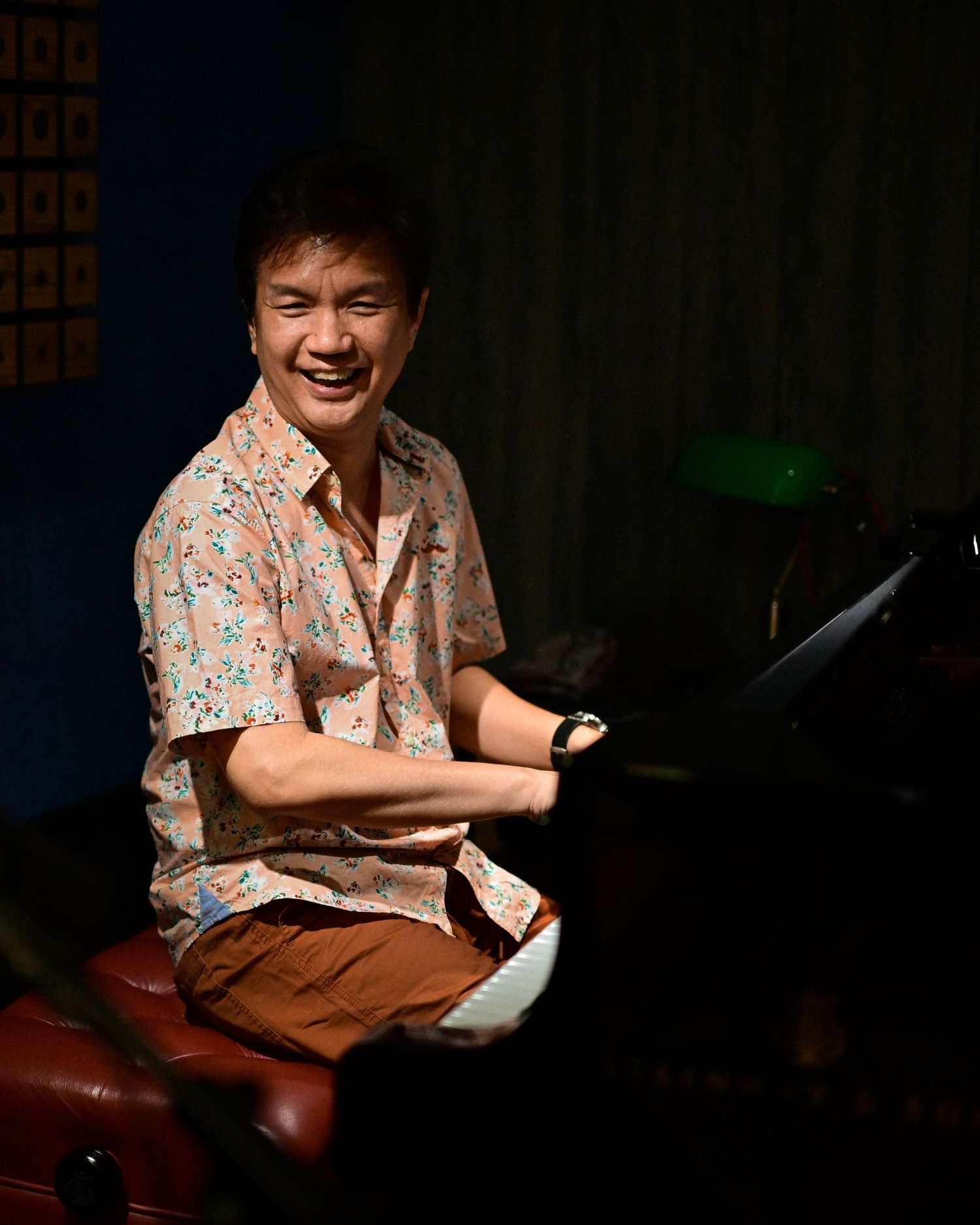 Smiling Asian man in floral shirt sits at a grand piano in a dimly lit music studio.