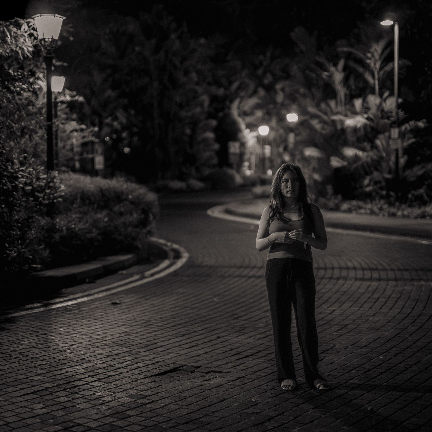 A woman stands alone on a brick road at night, illuminated by street lamps in a dark, tree-lined park.