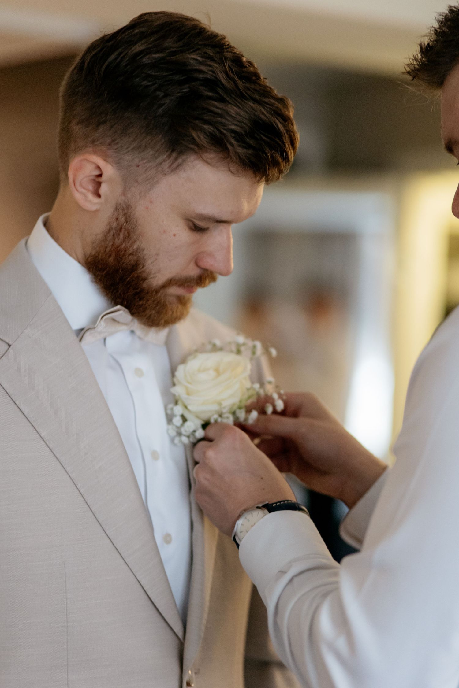 Groom in beige suit having white rose boutonniere pinned to lapel by groomsman indoors before wedding ceremony.