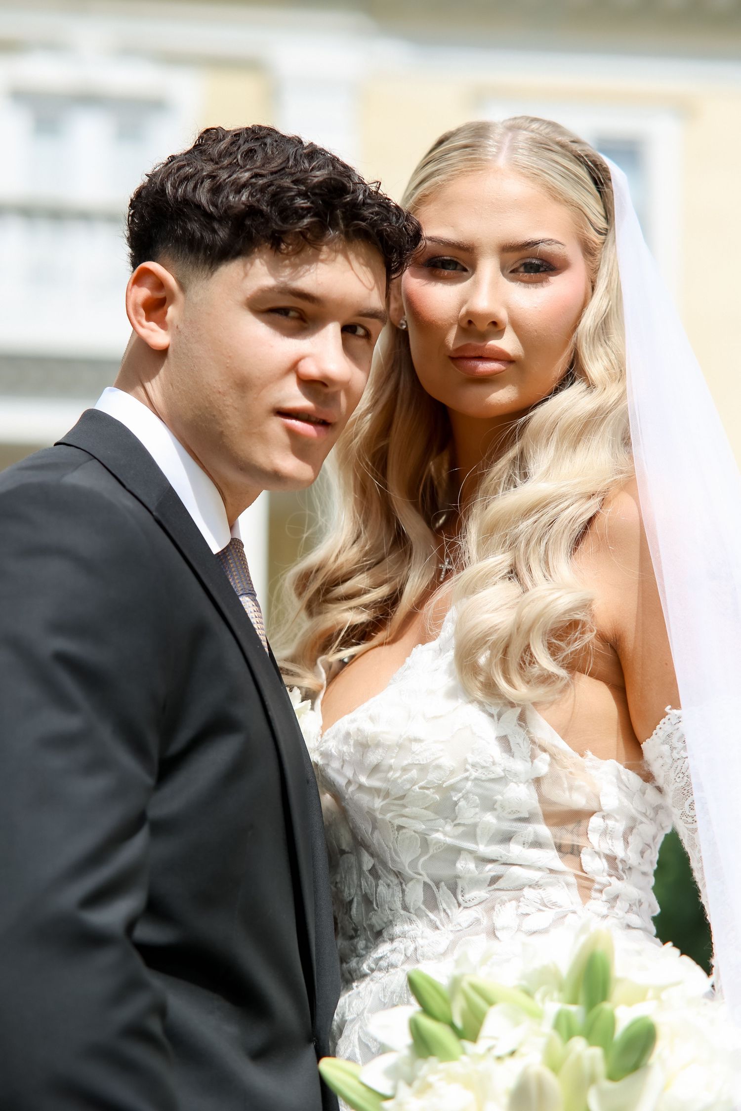 Young bride and groom posing together outdoors, she in white lace gown with veil, he in black suit.
