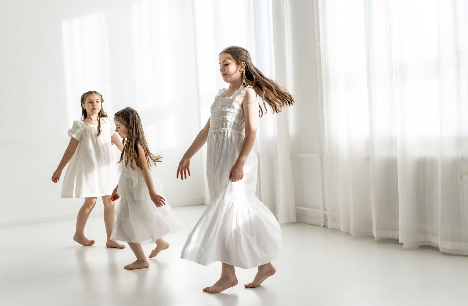 Three girls in white dresses dancing barefoot in a bright white room.