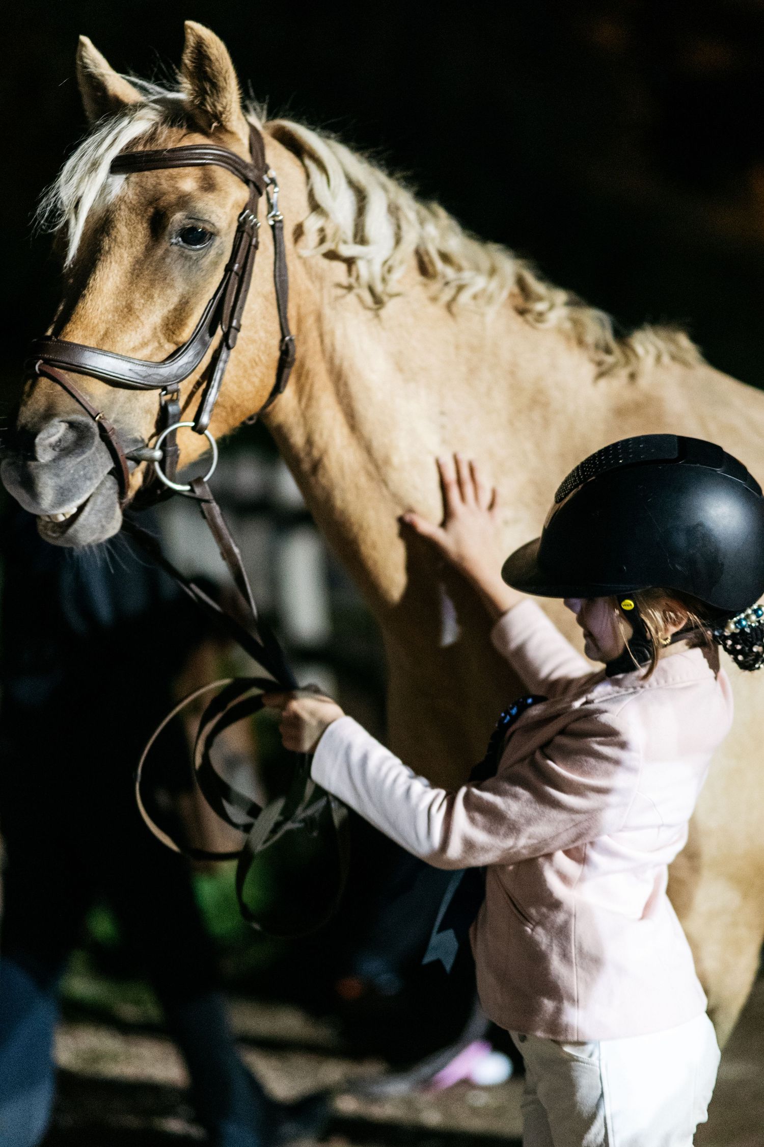 Young girl in riding helmet gently touches a palomino horse wearing a bridle in a stable.