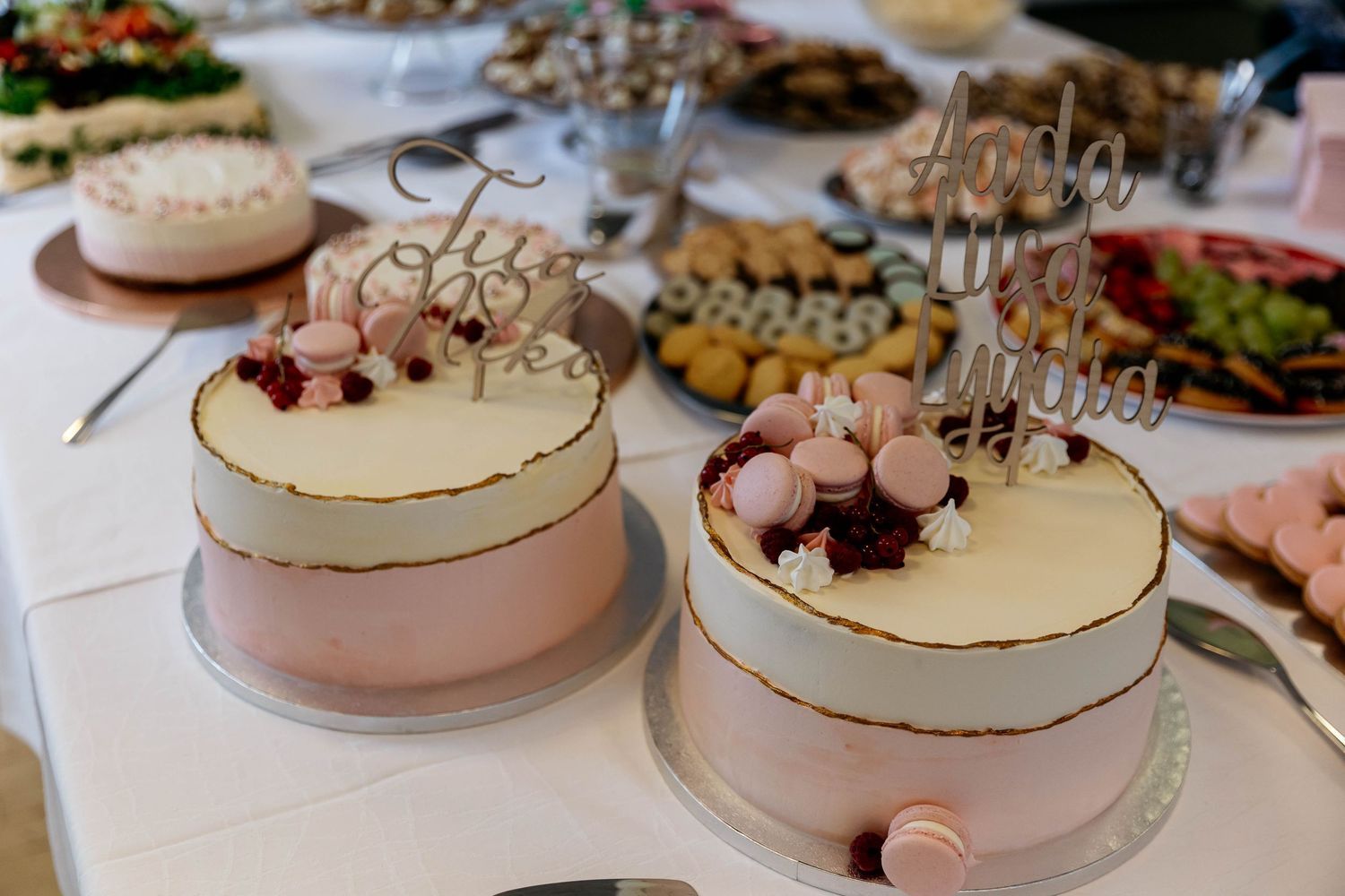 Two elegant white and pink celebration cakes with gold trim and decorative toppers on a dessert table.