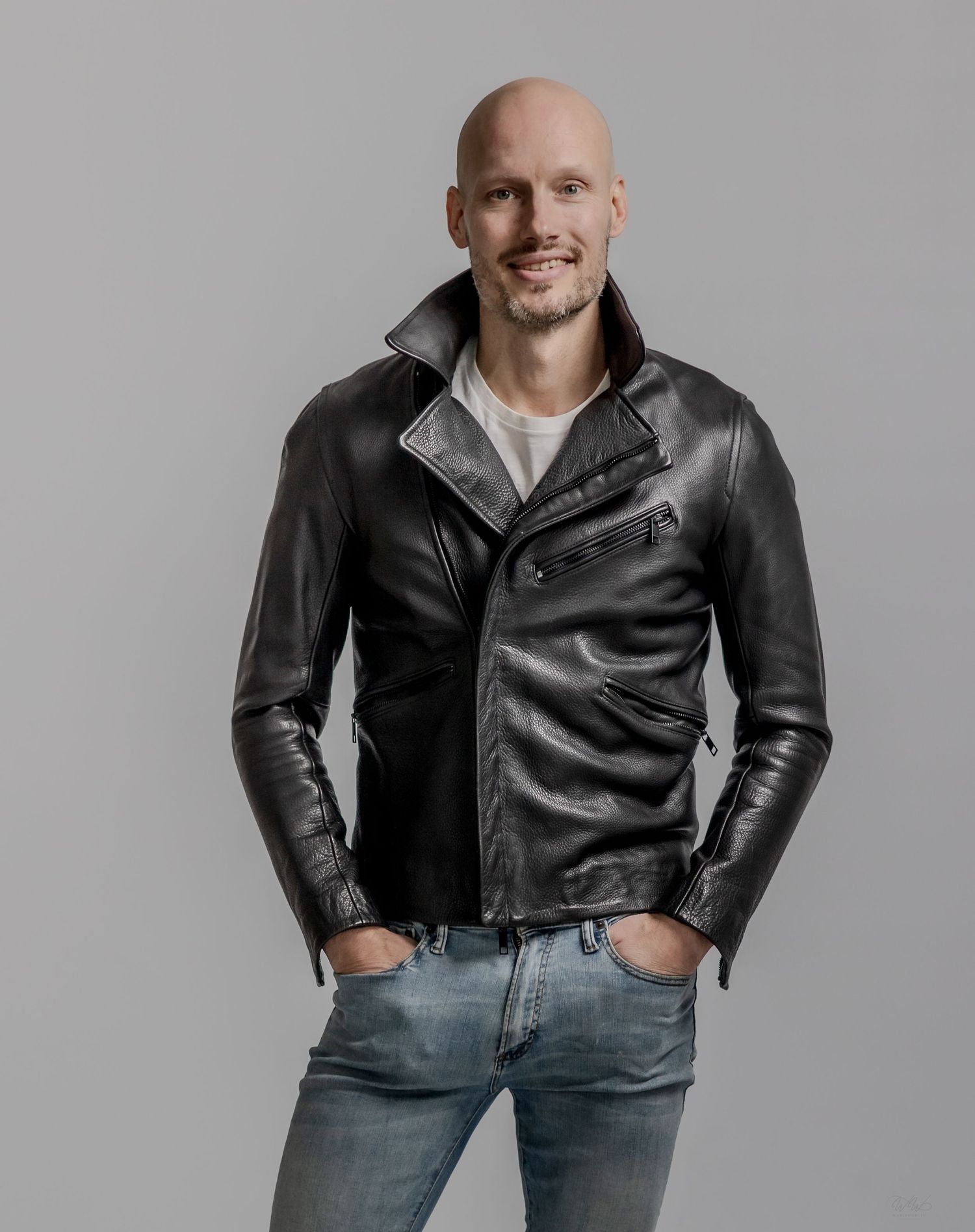 Bald man smiling in black leather biker jacket and jeans posing with hands in pockets against grey background.