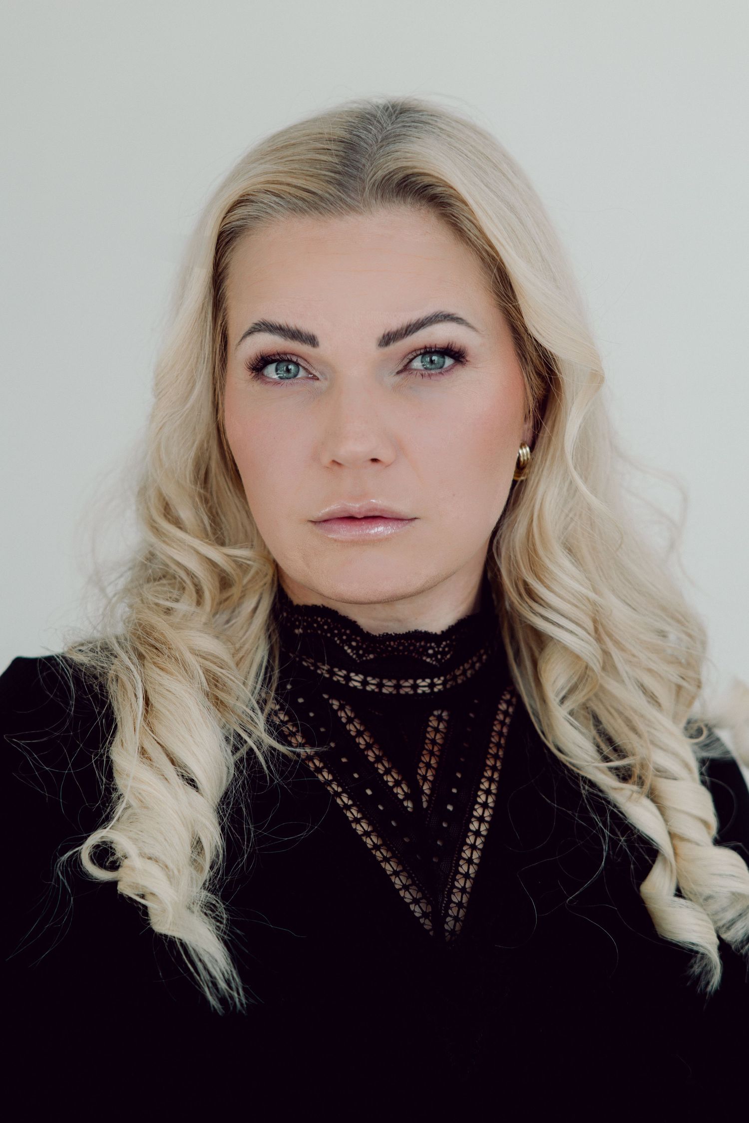 Portrait of a woman with long wavy blonde hair, blue eyes and a black lace-trimmed high-neck top against a plain light background.