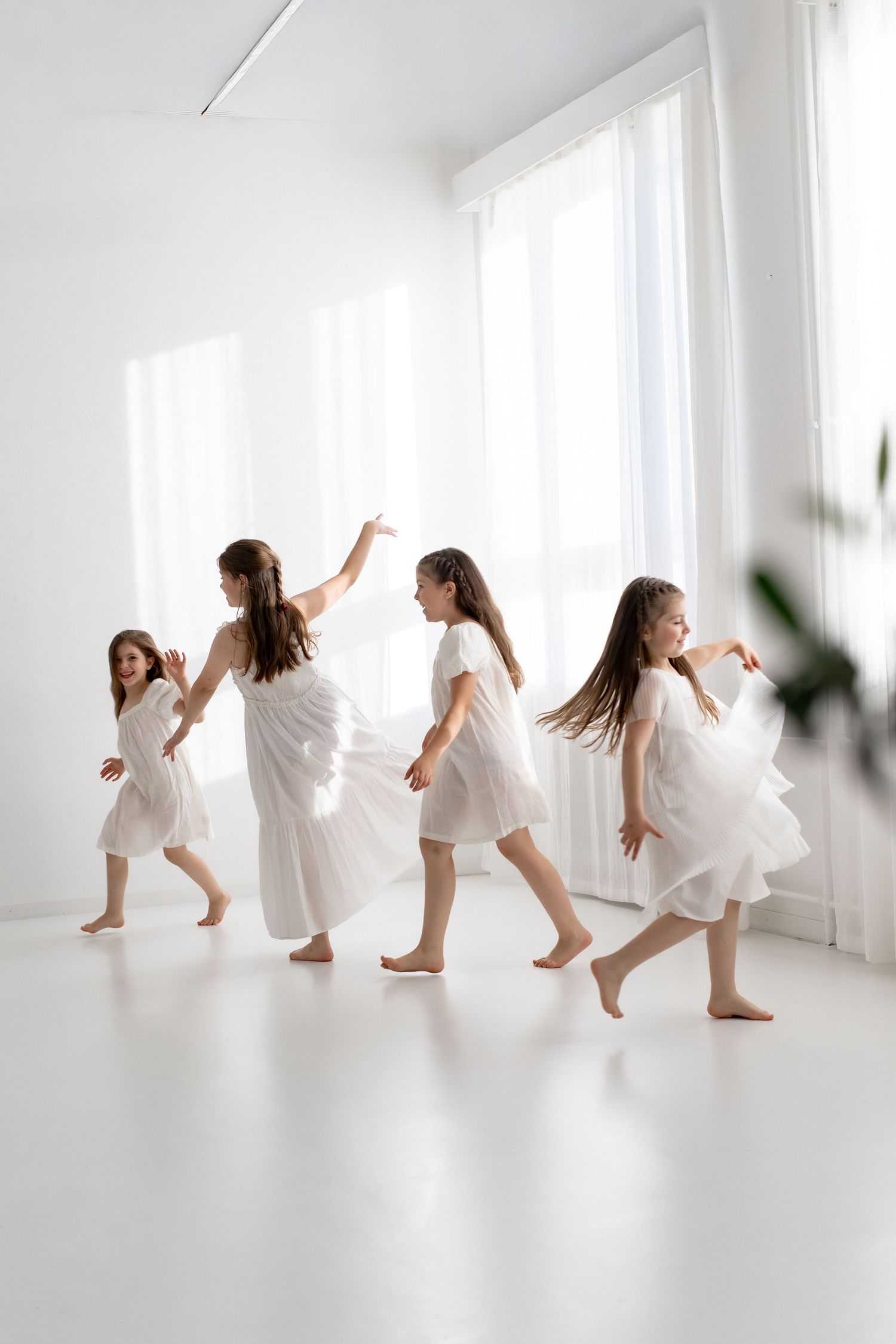 Four girls in white dresses dancing barefoot in a bright white studio with natural light from large windows.