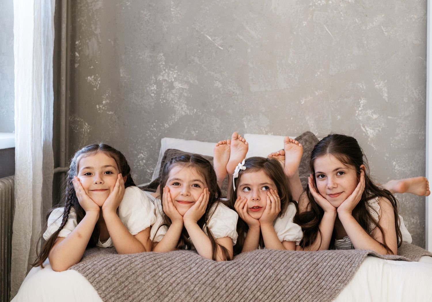 Four young girls lying on a bed smiling and posing together in matching white outfits in a cozy bedroom.