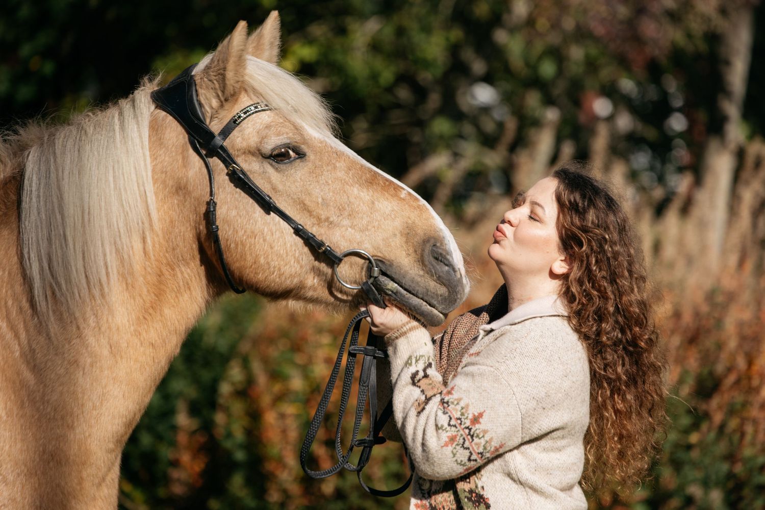 A young woman with curly hair gently bonds with a palomino horse outdoors in warm golden sunlight.
