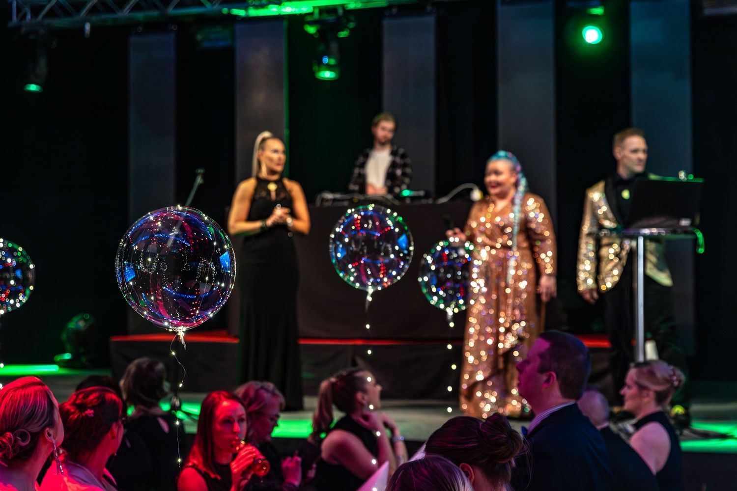Colorful gala event stage with performers in sequined outfits and LED balloons under green stage lighting.