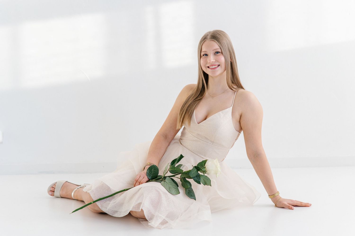 Smiling young woman with long hair sits on a white floor in a flowing blush dress, holding a single green-leafed rose stem.