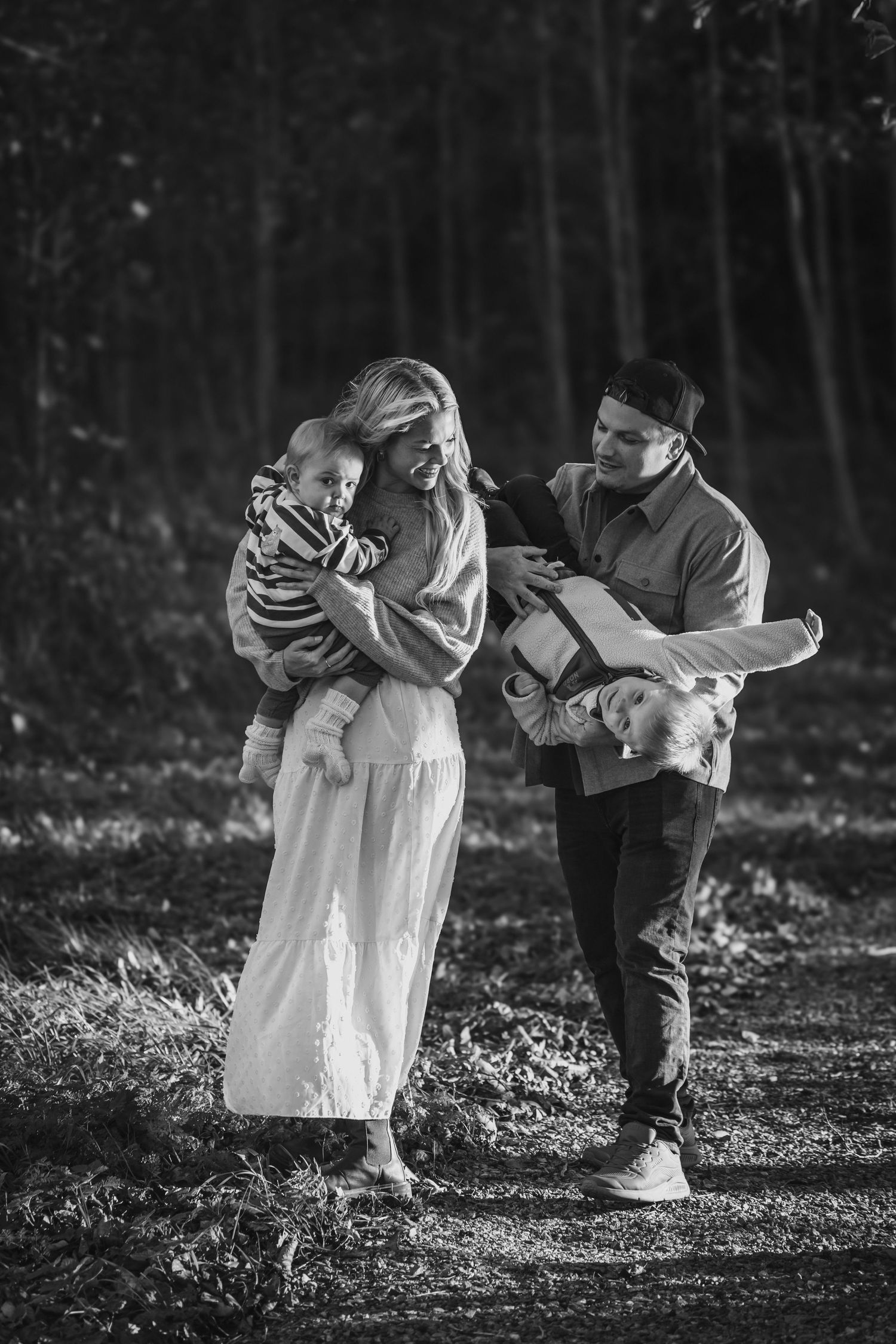 Black and white photo of a smiling family of four walking on a wooded path, with a mother and father carrying their two young children.