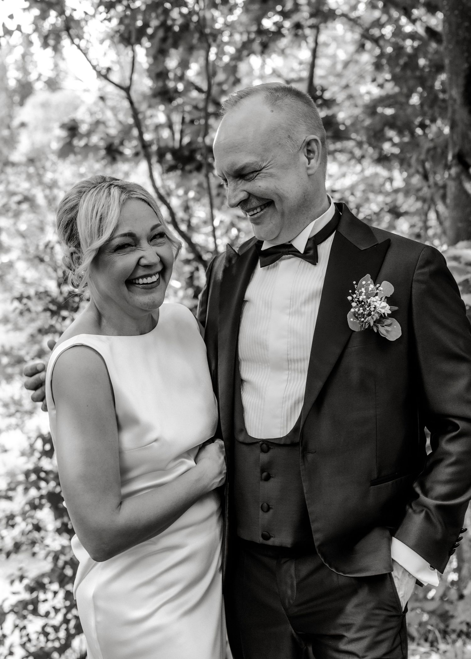 Black and white photo of laughing bride and groom in tuxedo embracing outdoors among trees.