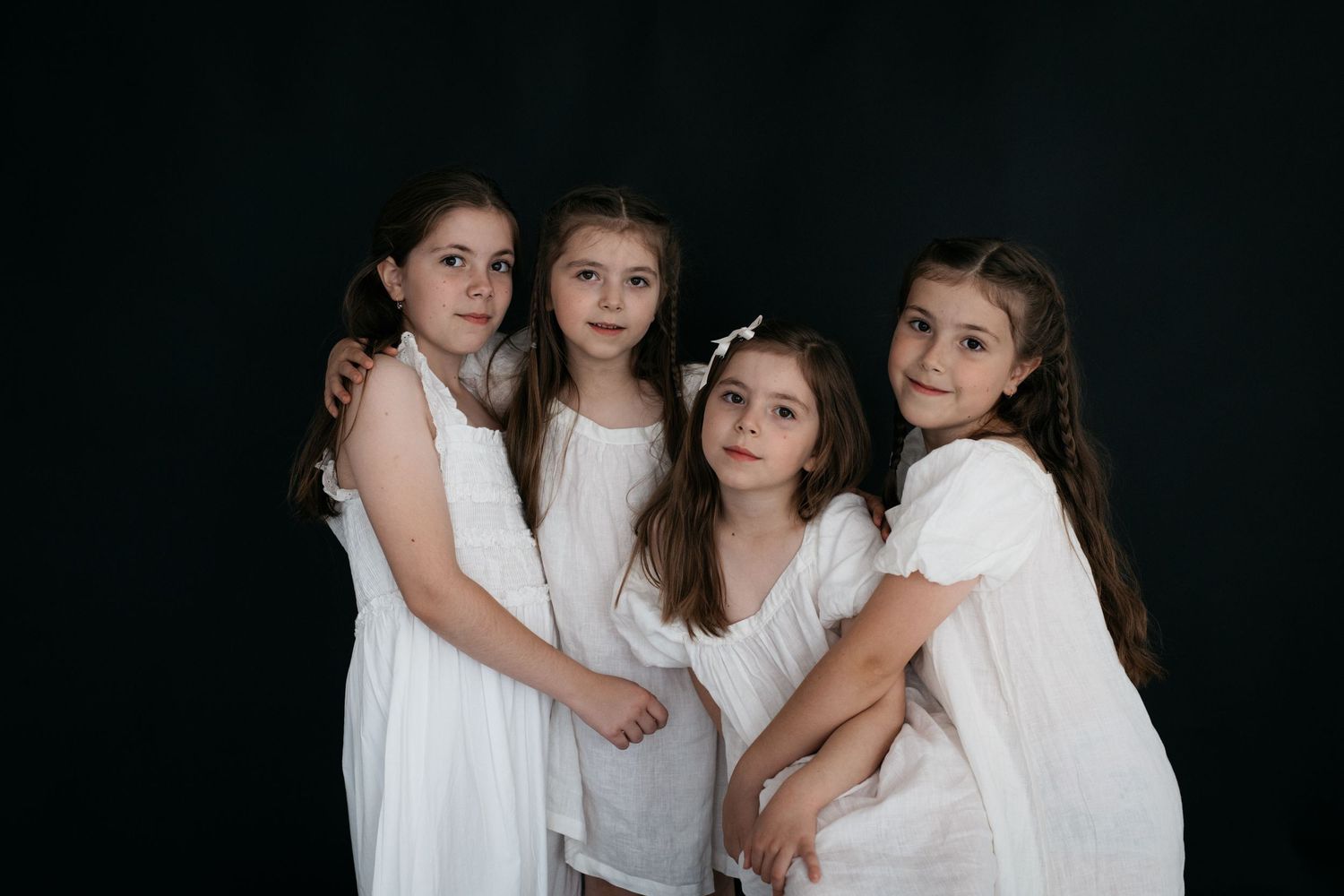 Four young girls in white dresses pose together against a dark background in a studio portrait.