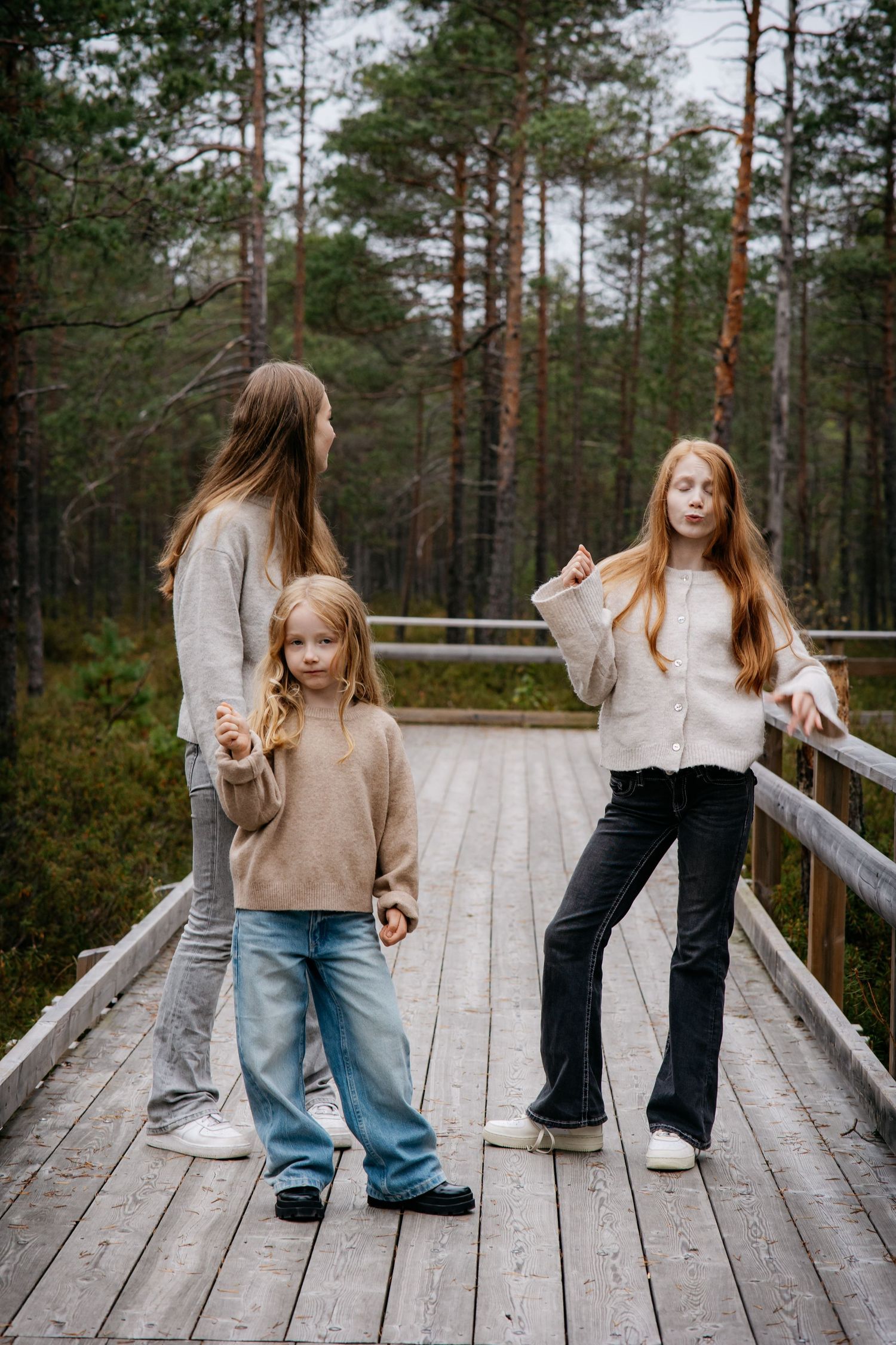 Three girls standing on a wooden bridge in a forest, wearing casual sweaters and jeans on a cloudy day.