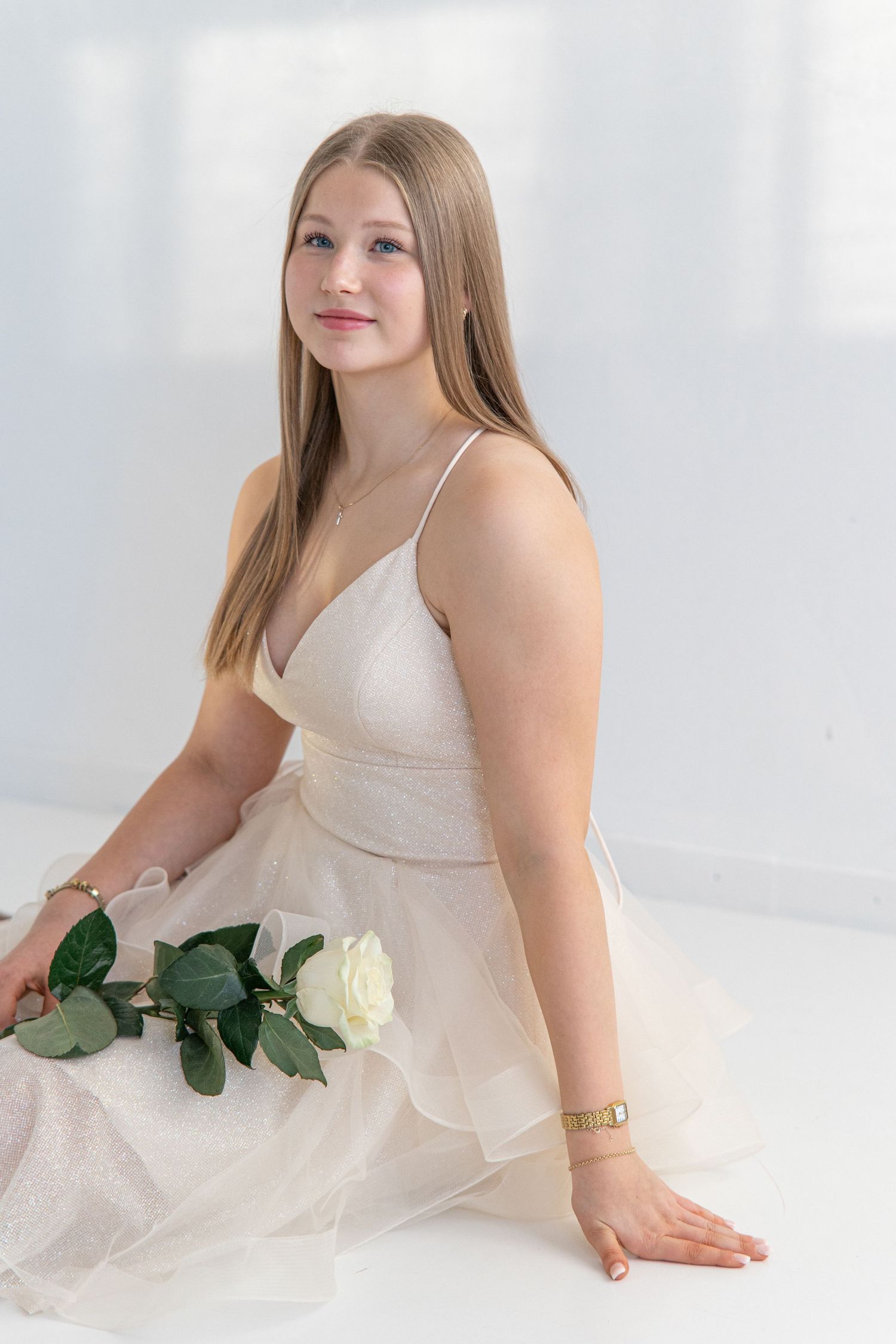 Young woman with long straight hair sits on a white floor in a champagne tulle gown, holding a white rose with green leaves.