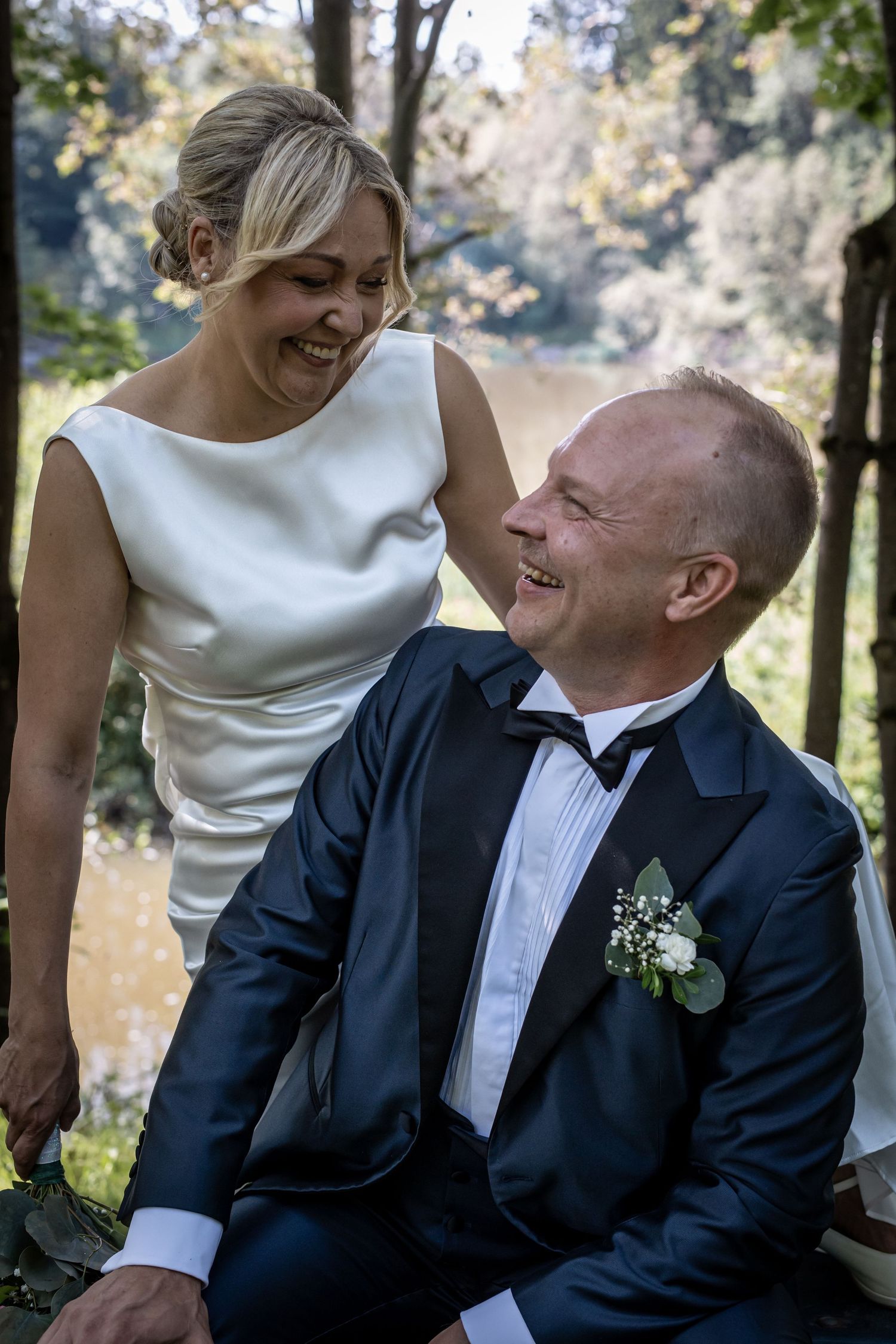 Bride and groom laughing together while seated outdoors near a sunlit pond surrounded by trees.