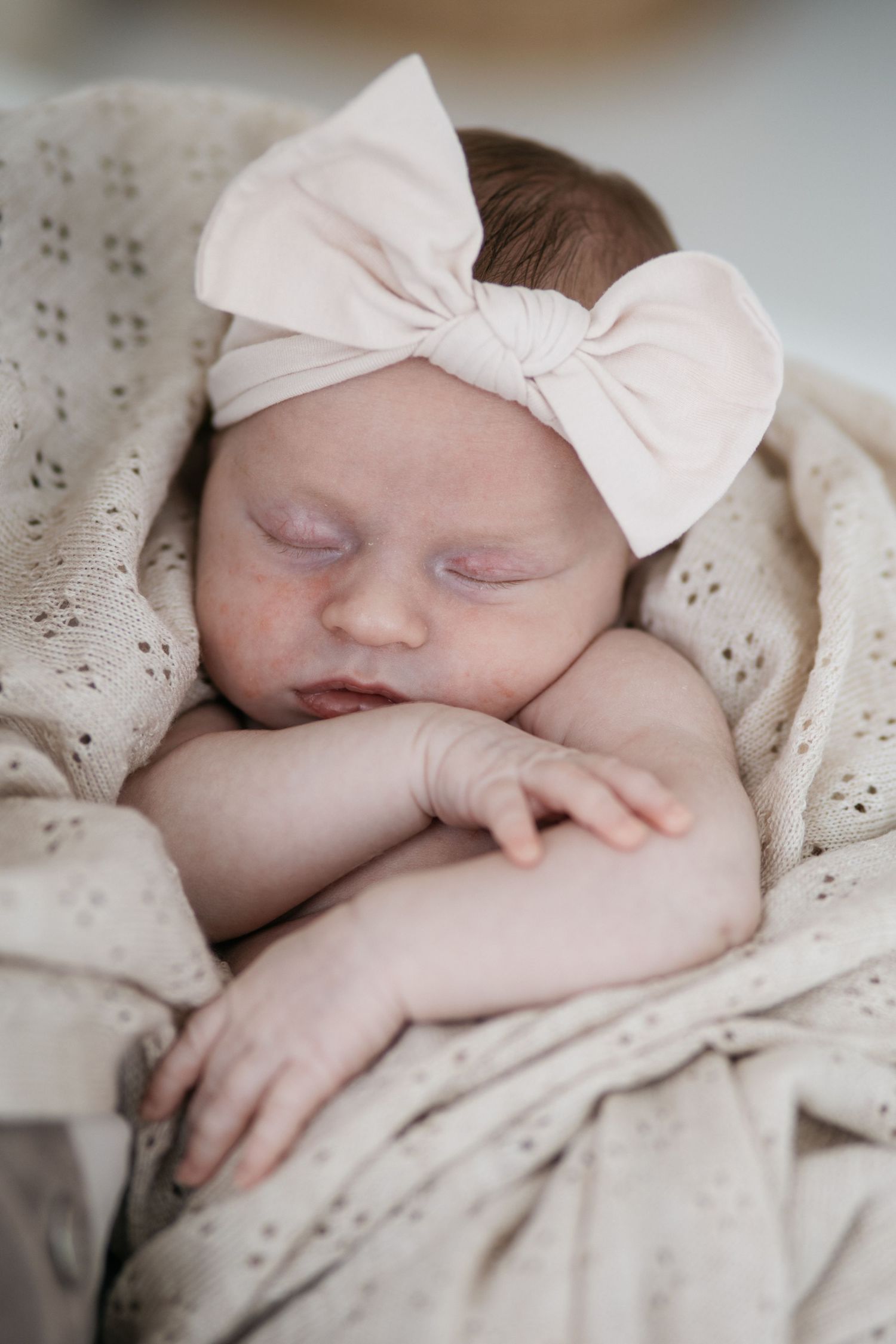 Sleeping newborn baby girl wearing a pink bow headband, wrapped in a soft beige knit blanket.
