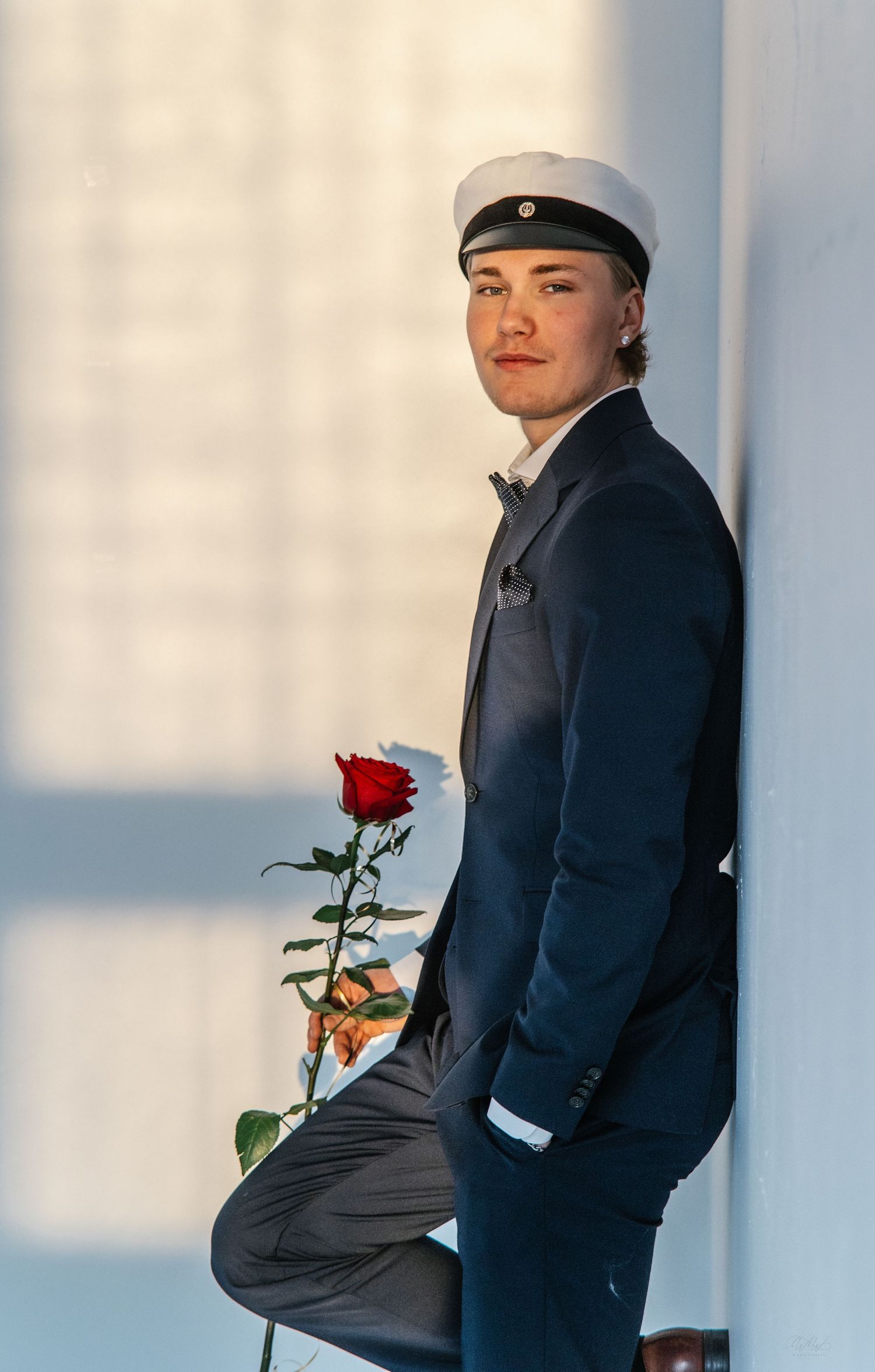 Young man in a dark suit and white student cap leans against a wall, holding a single red rose in soft natural light.