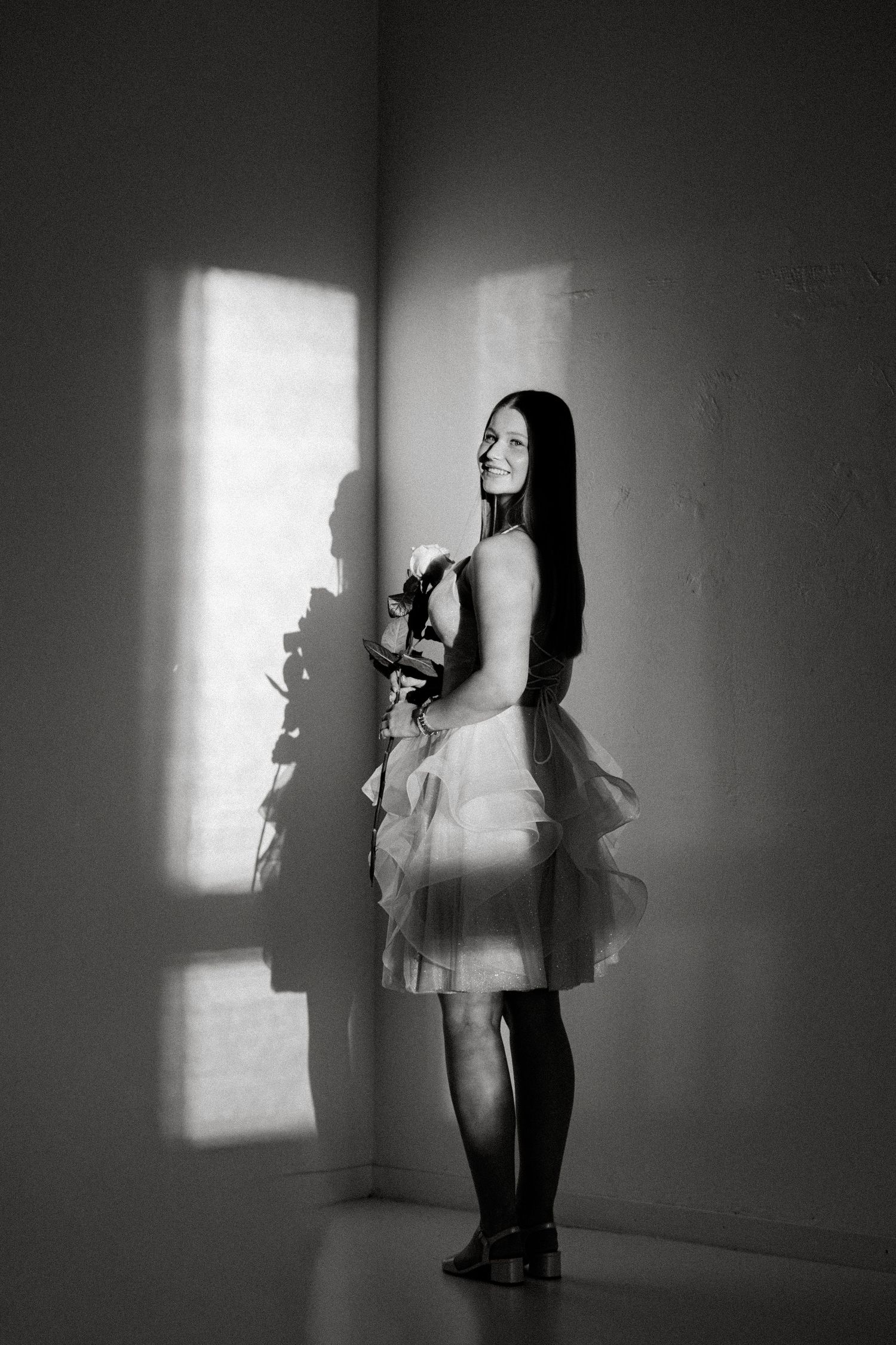 Black and white portrait of a smiling woman in a tulle skirt holding a rose, standing in a corner with window light and shadows.