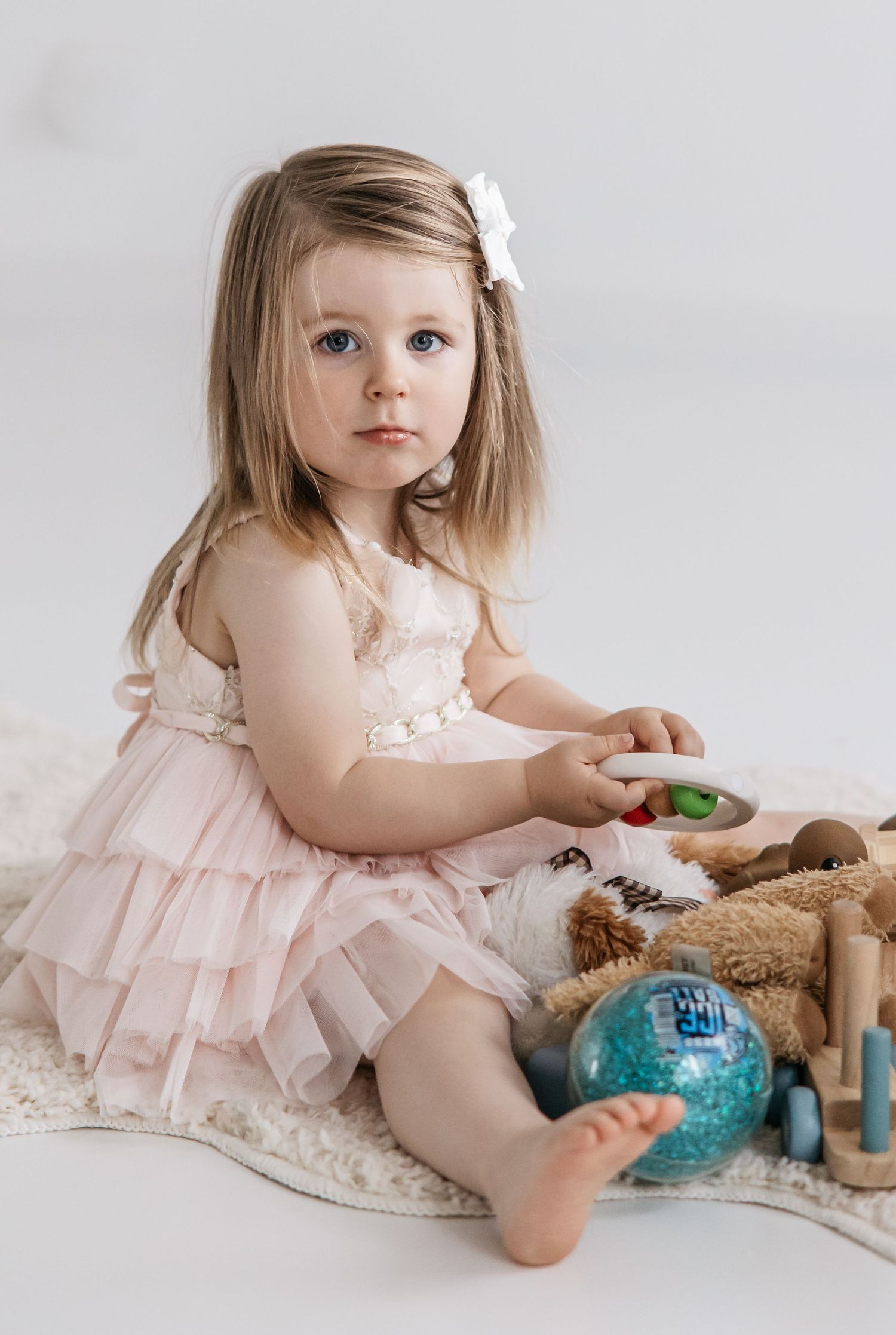 Toddler girl in a pink tulle dress with a white hair bow sits on a cream rug playing with wooden toys and a blue sensory ball.