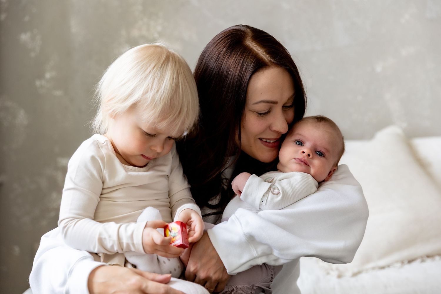 A mother with dark hair holds a newborn baby while her blonde toddler examines a small toy together.