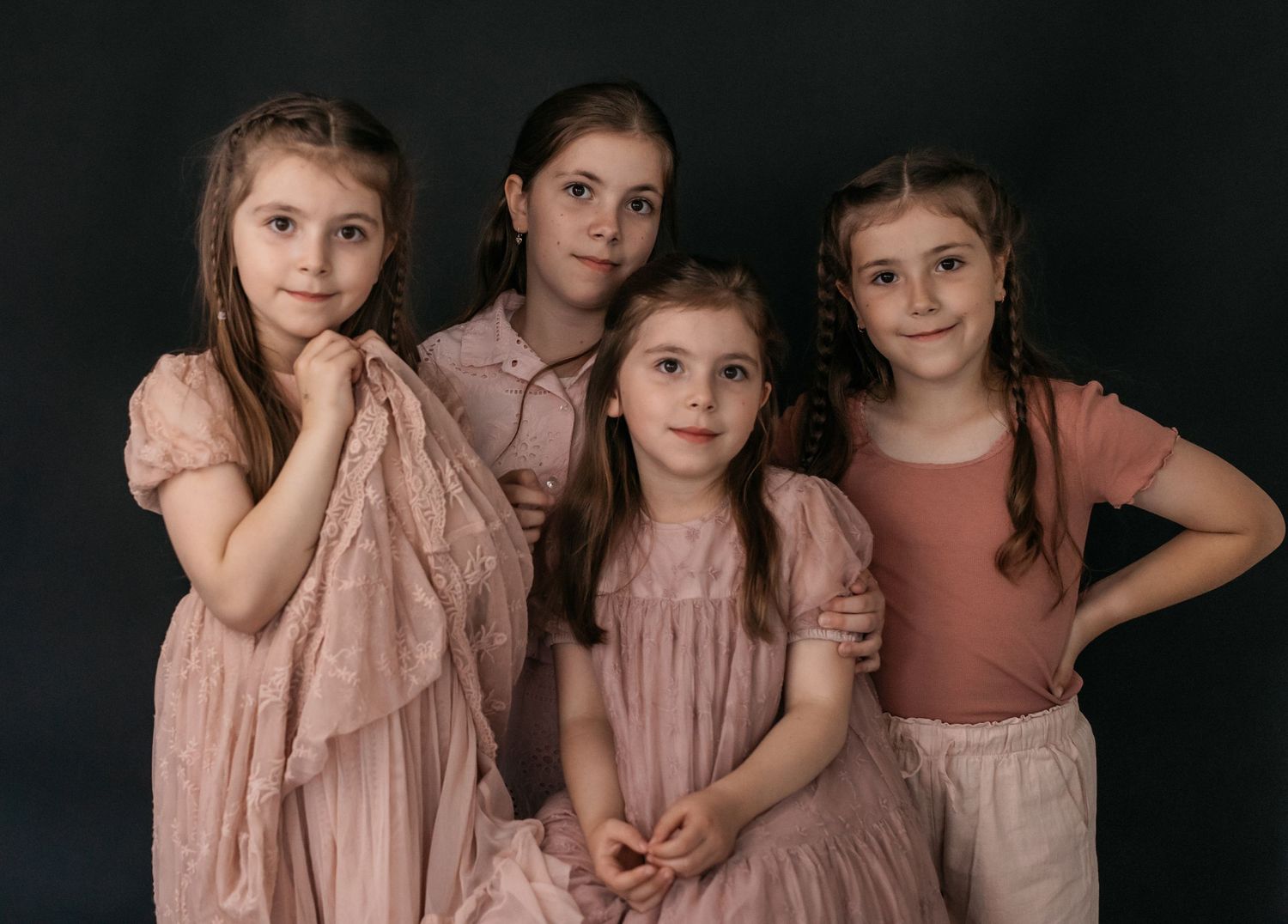 Four young girls in matching pink dresses pose together for a professional studio portrait against a dark background.