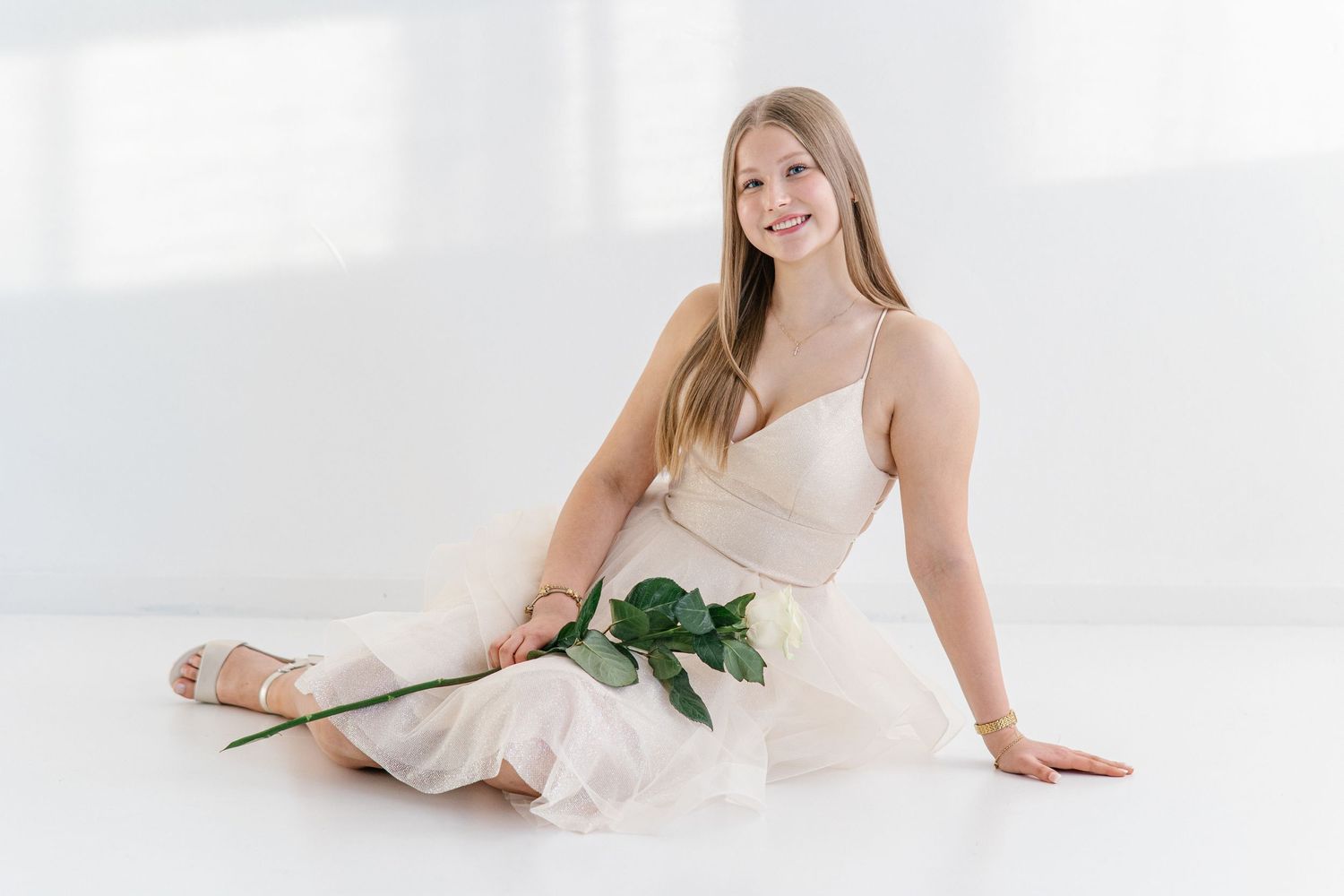 Smiling young woman with long blonde hair sits on a white floor in a flowing blush dress, holding a single green-stemmed white rose.