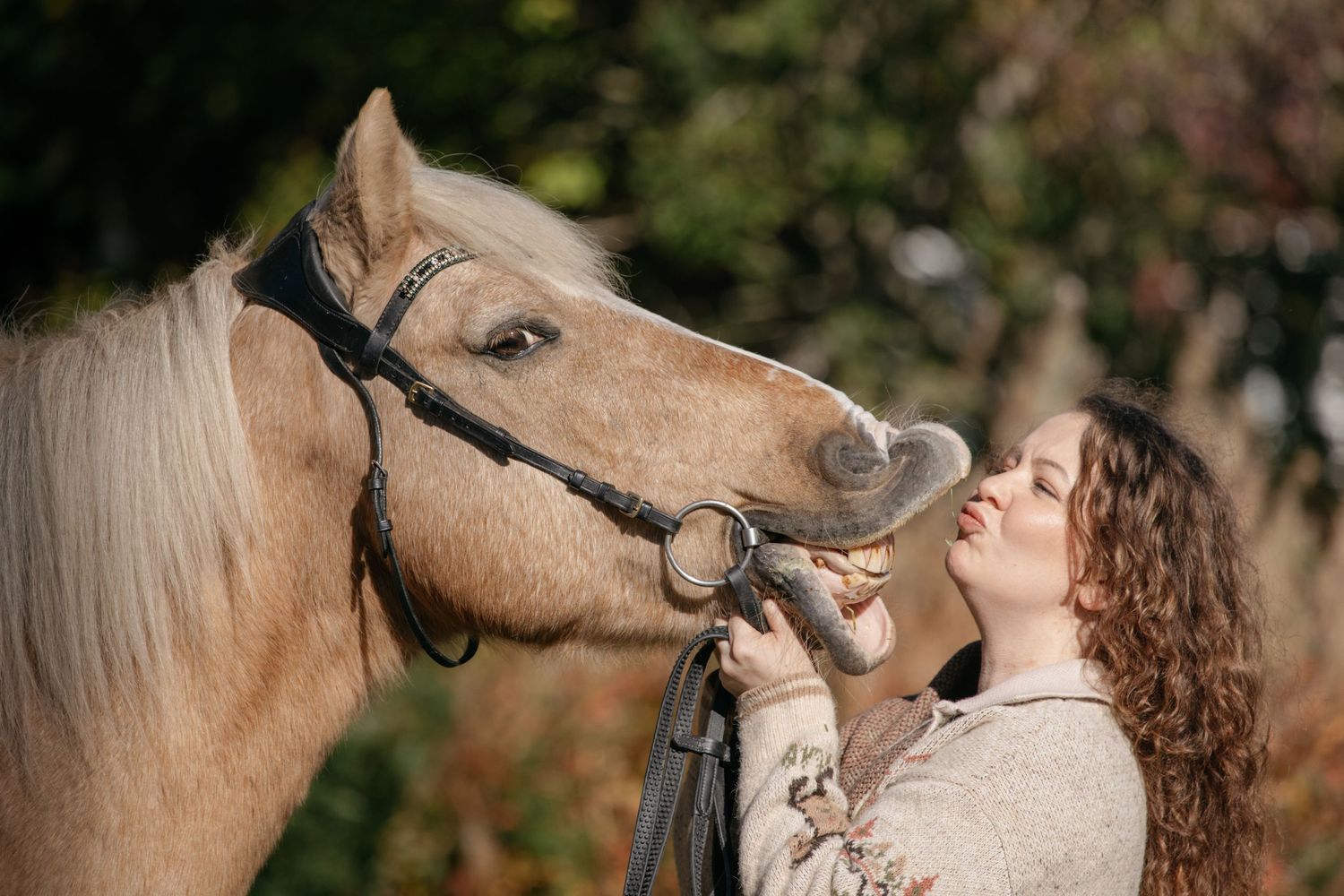 Young woman with curly hair smiling and bonding with a palomino horse wearing a halter outdoors.