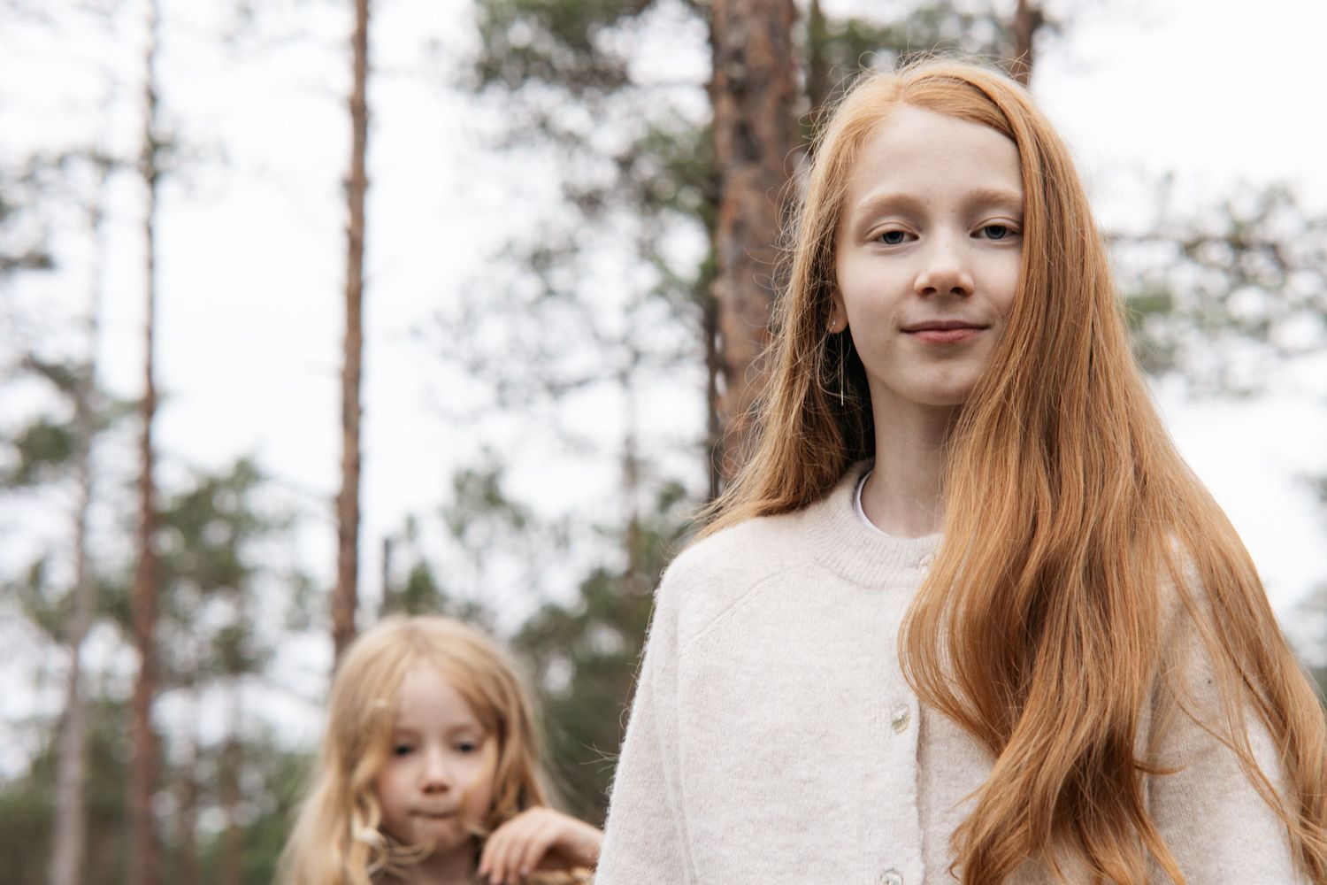 Two young redhead girls standing outdoors in a pine forest, with the older girl smiling at the camera.