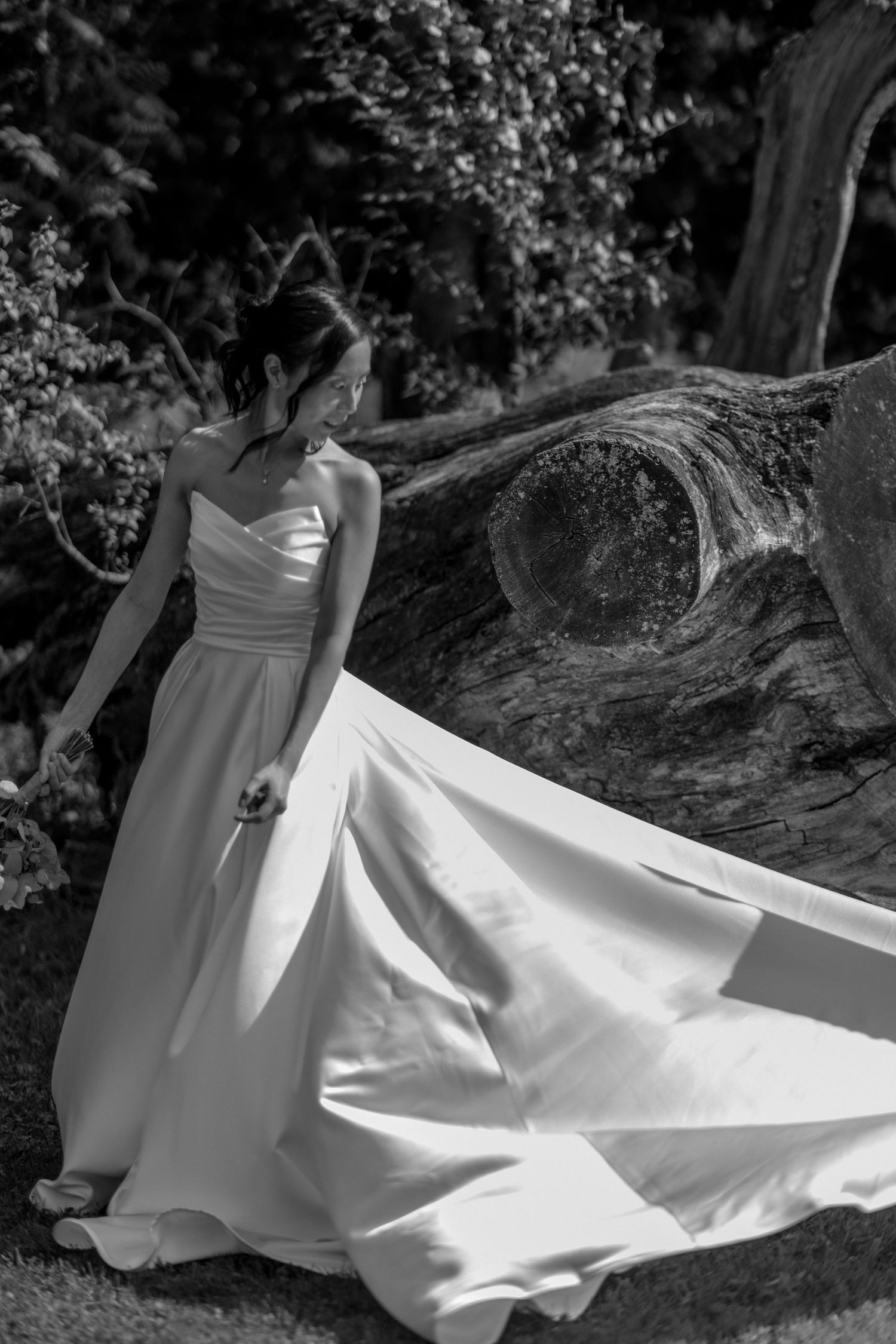 Elegant bride in flowing white strapless gown posing near a large tree trunk in black and white photo.