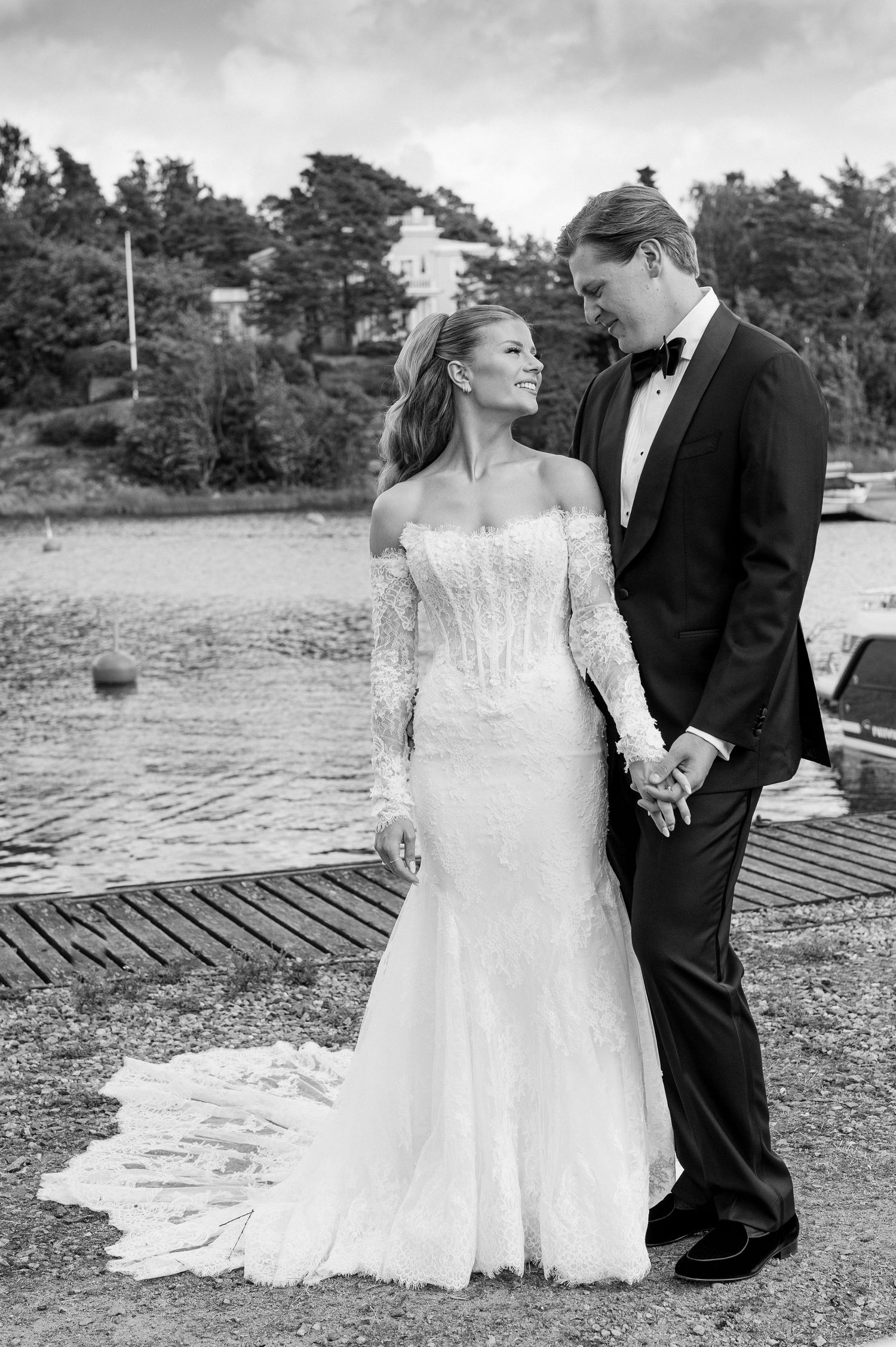 Elegant black and white wedding photo of a bride in a lace off-shoulder gown and groom in a tuxedo by the waterfront.