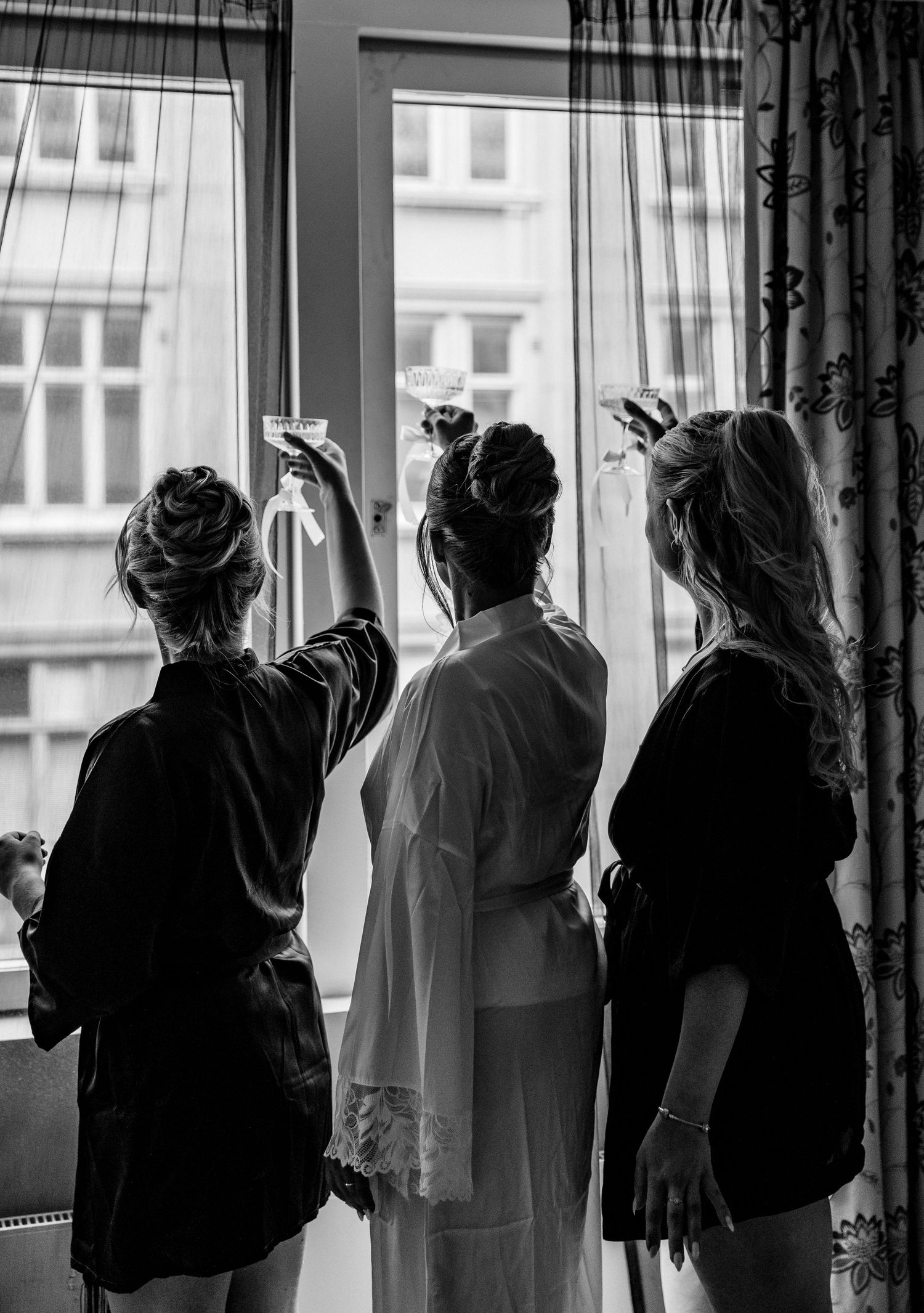 Black and white photo of three women in robes raising glasses by a window in a bridal getting ready moment.