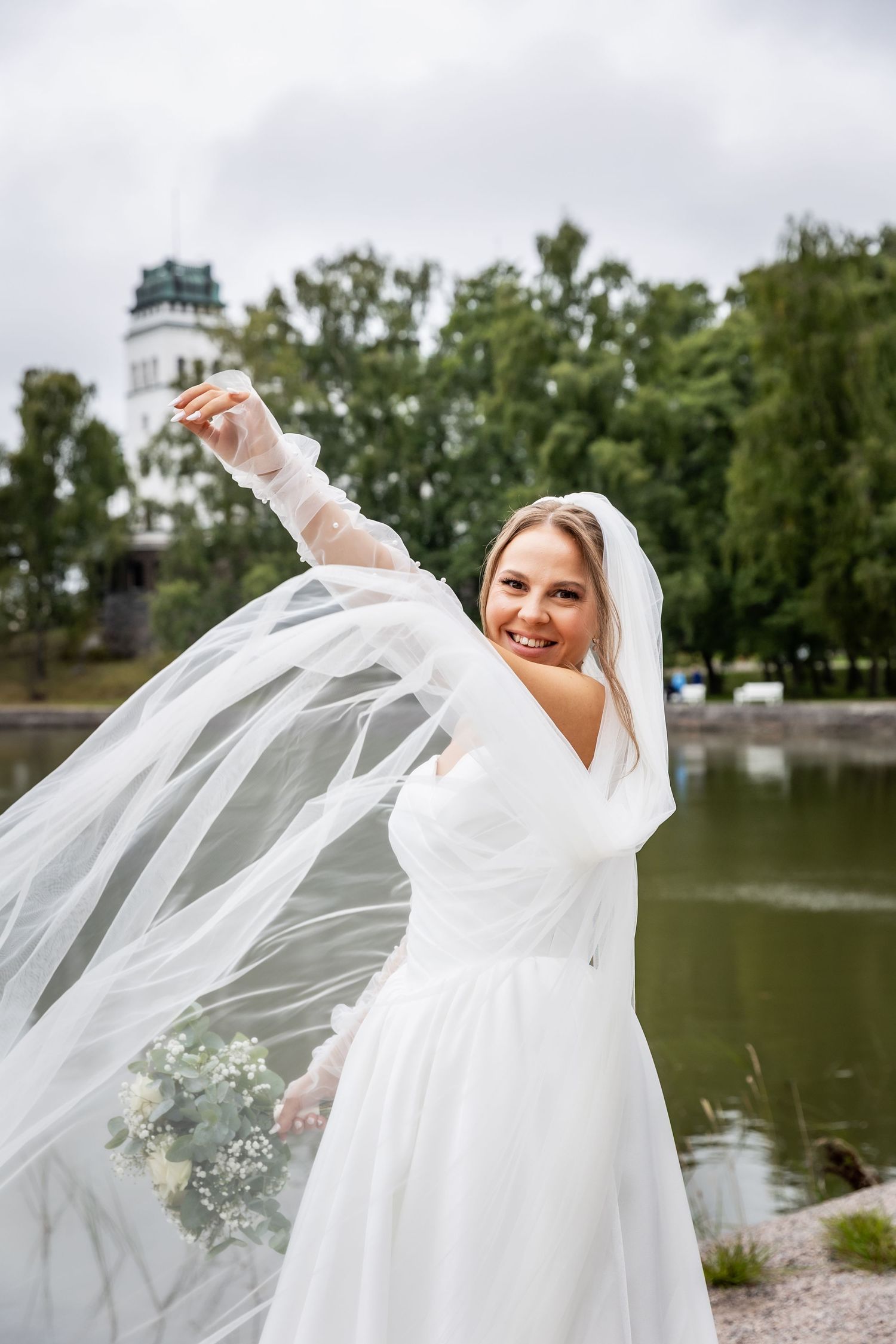 Smiling bride in white gown and veil twirling outdoors near a lake with green trees and a tower in background.