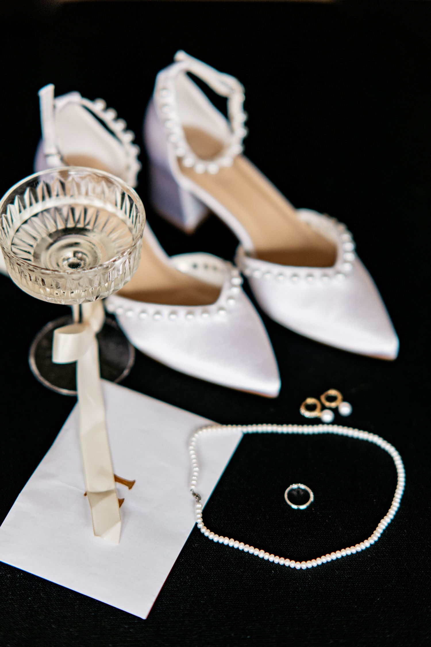 White pearl-trimmed bridal heels displayed with pearl necklace, gold earrings, ring, and crystal glass.