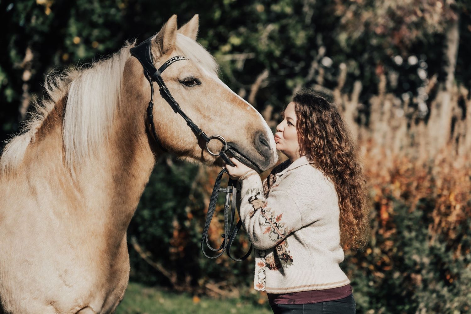 Young woman with curly hair tenderly bonding with a palomino horse wearing a bridle outdoors.