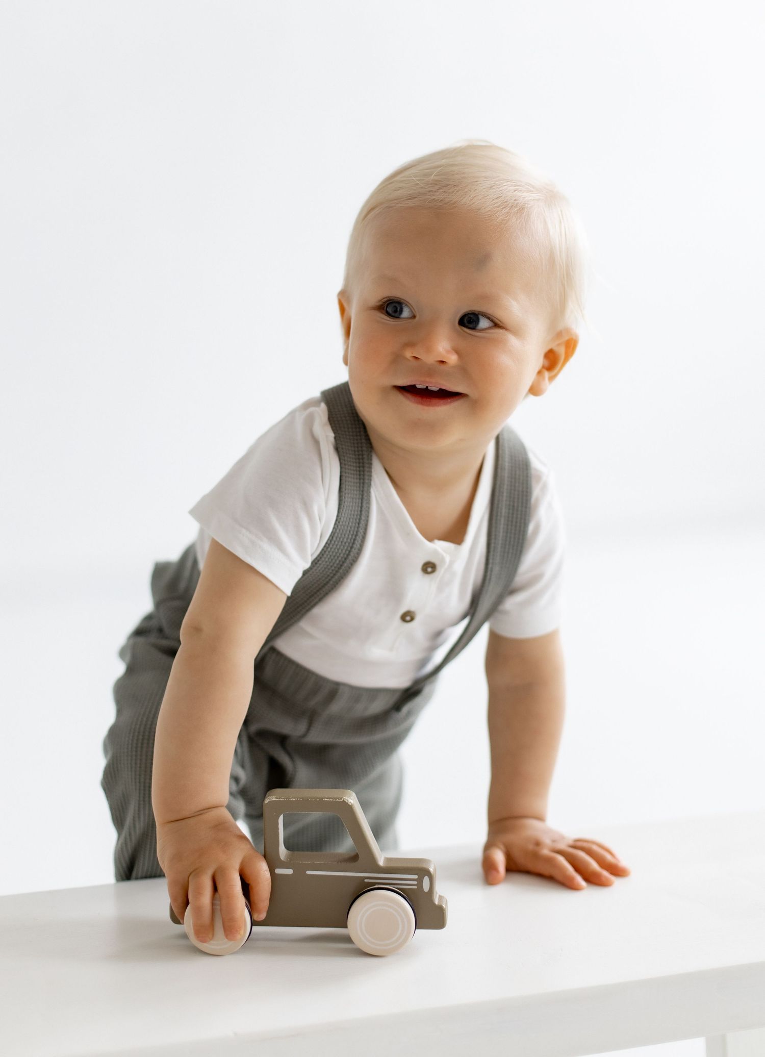 Smiling blond toddler in a white shirt and gray suspender pants leans on a white bench while playing with a wooden toy tractor.