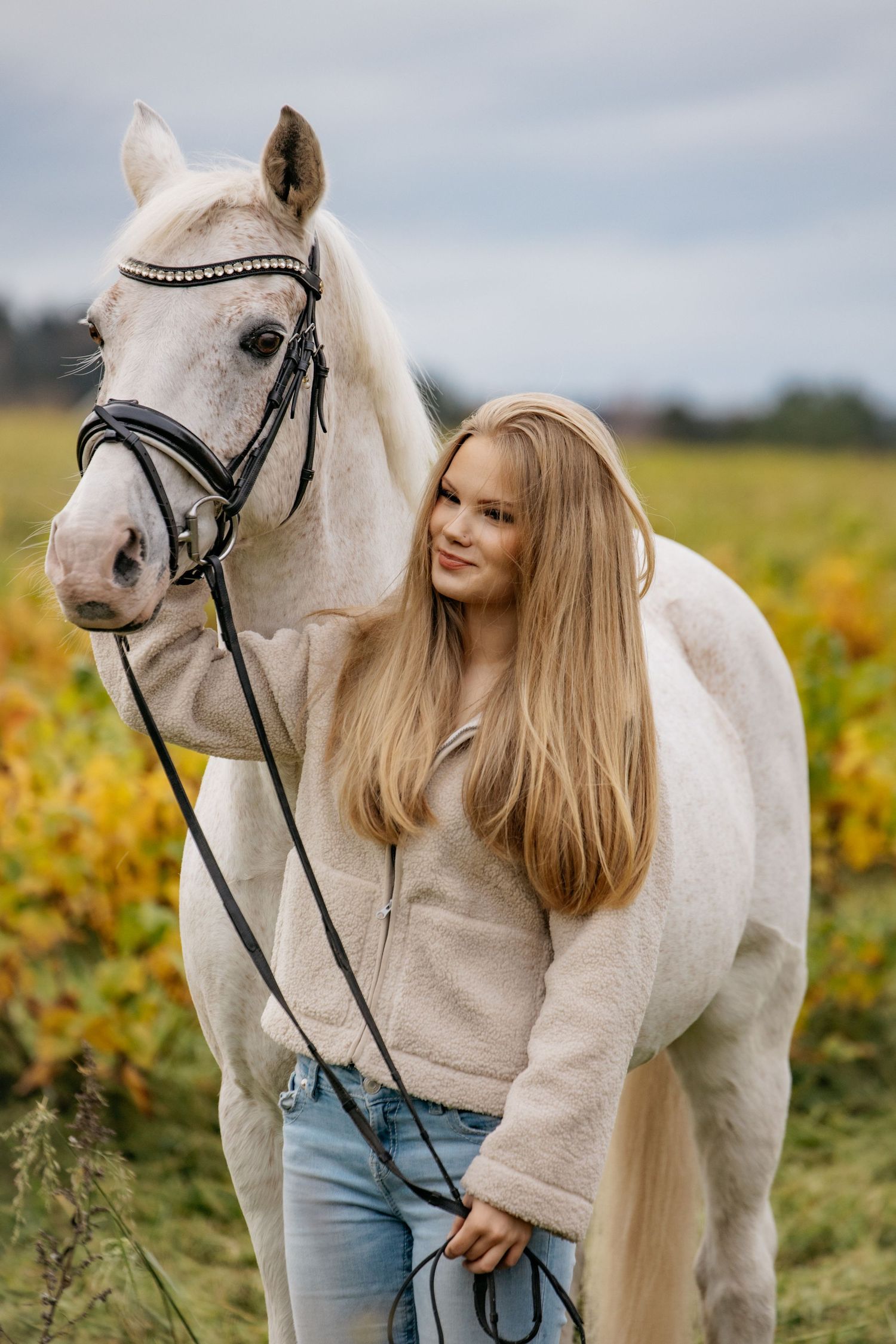 Young blonde woman in beige fleece jacket and jeans stands beside a white horse in an autumn field.