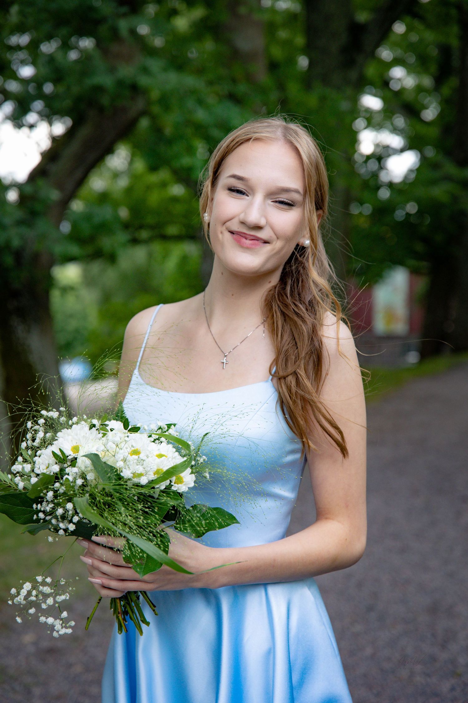 Smiling young woman with long strawberry-blonde hair in a light blue satin dress holding a white flower bouquet outdoors under green trees.