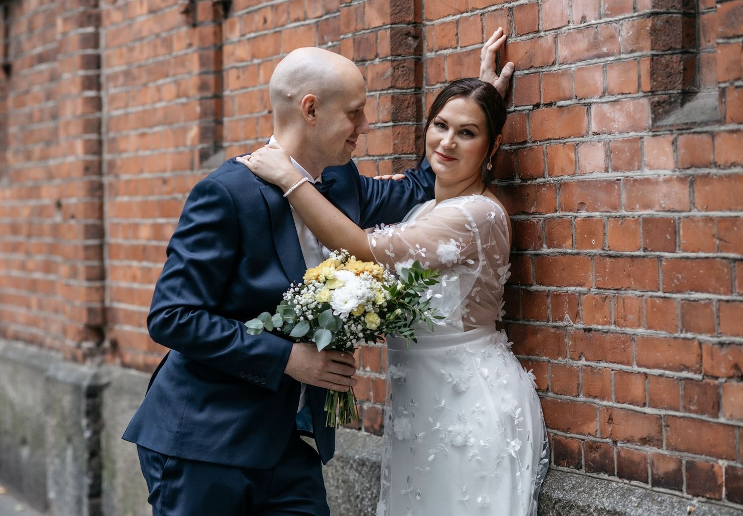 Bride and groom posing against a red brick wall, bride holding a bouquet of white and yellow flowers.