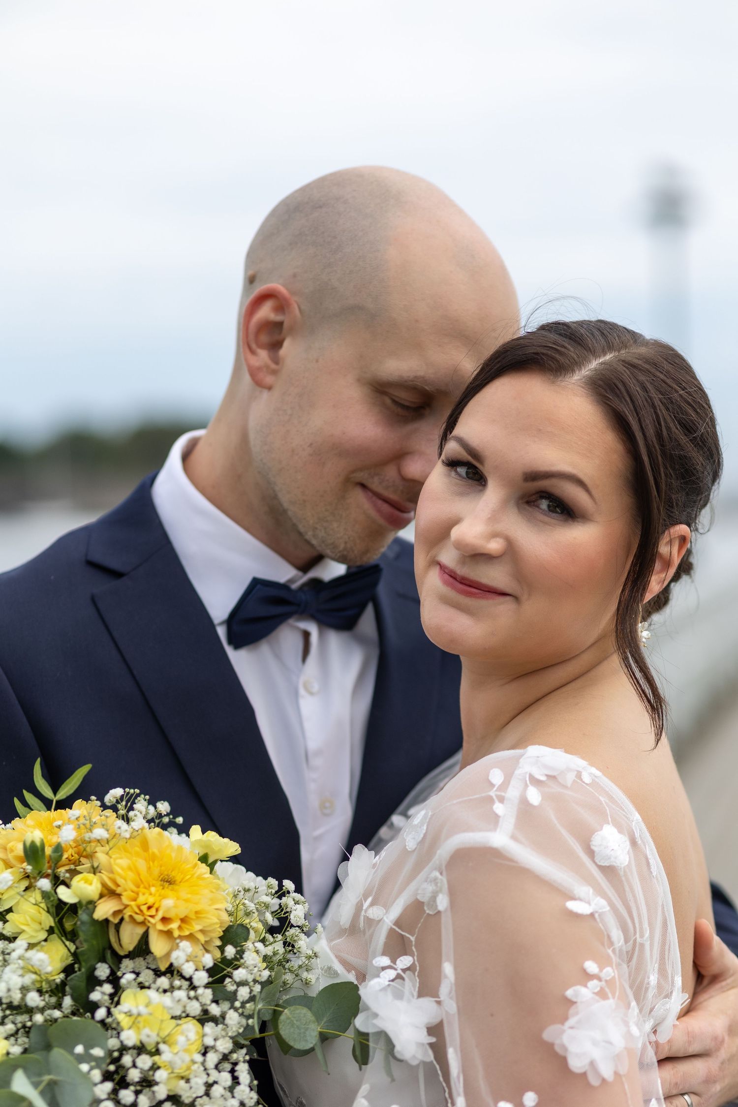 Bride holding yellow floral bouquet leans into bald groom in navy bow tie suit near waterfront.
