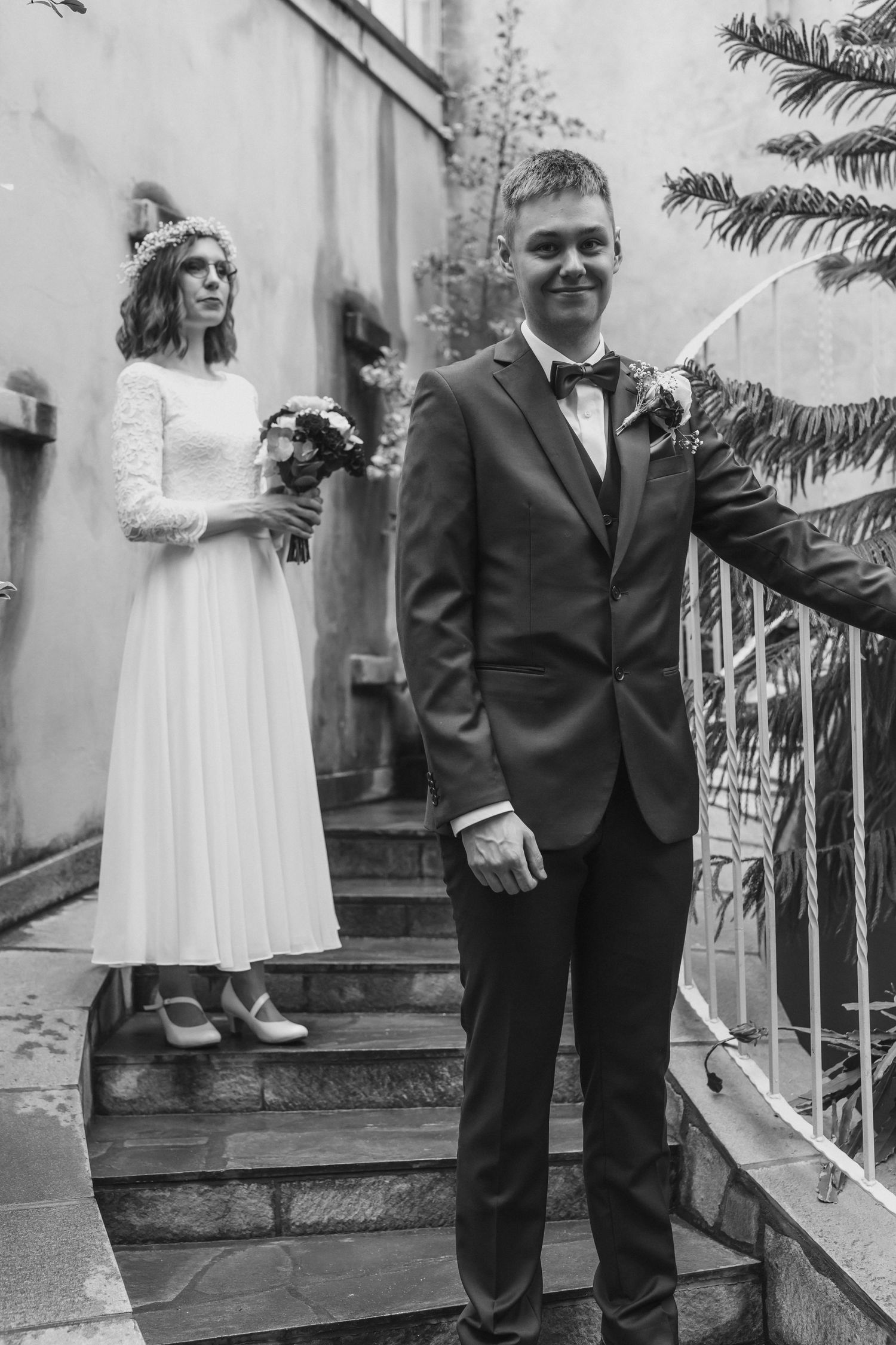 Black and white photo of groom in suit on steps with bride in white dress and flower crown behind him.