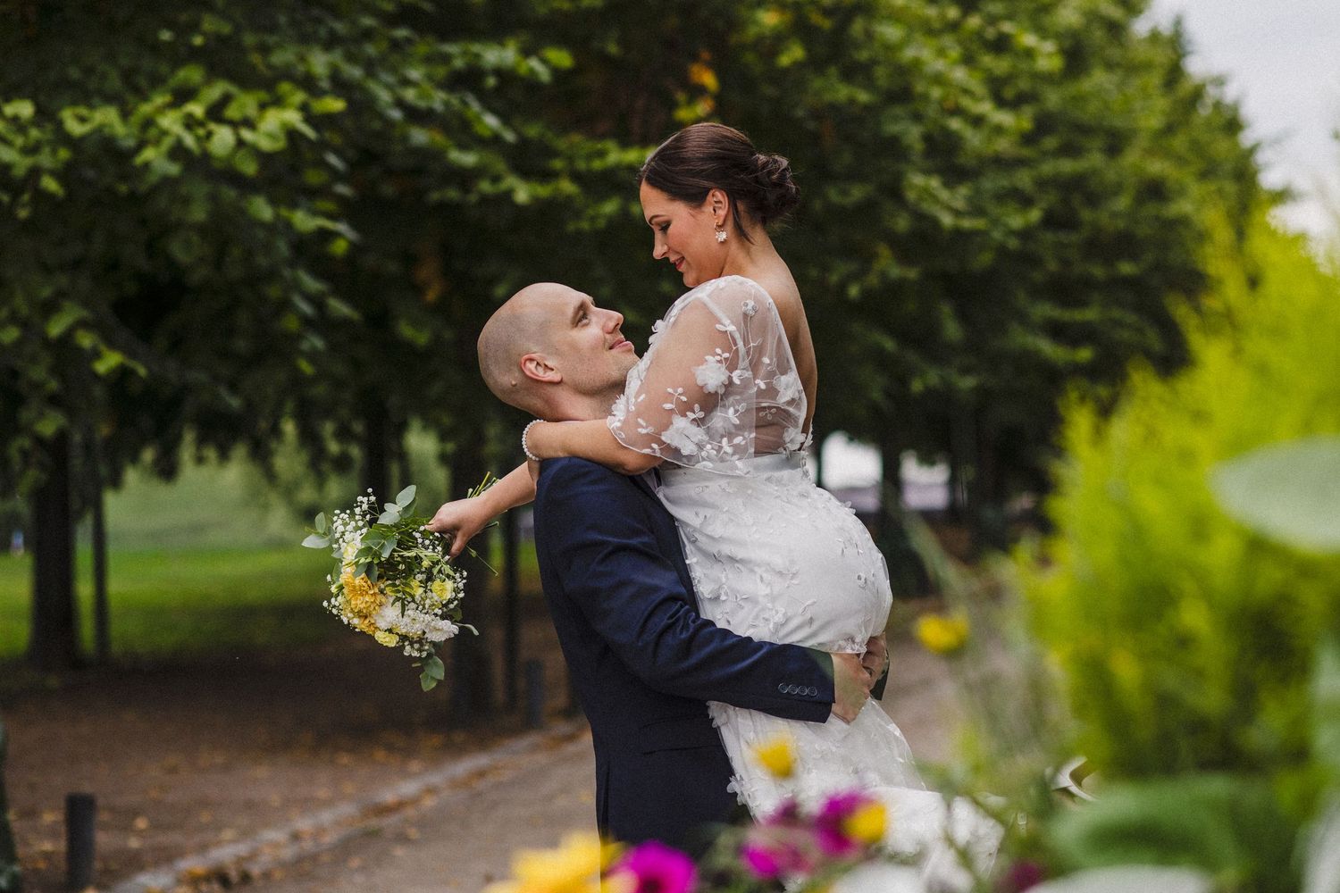 A groom lifts his bride in a lush green park, as she holds a colorful bouquet in her wedding dress.