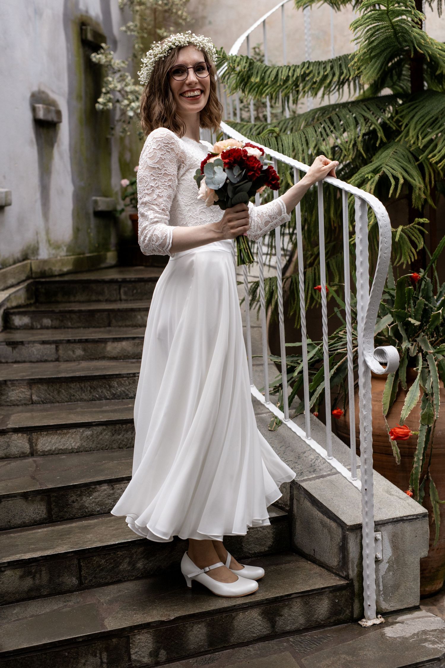 Smiling bride in white lace dress and flower crown holding red bouquet on stone steps with greenery.