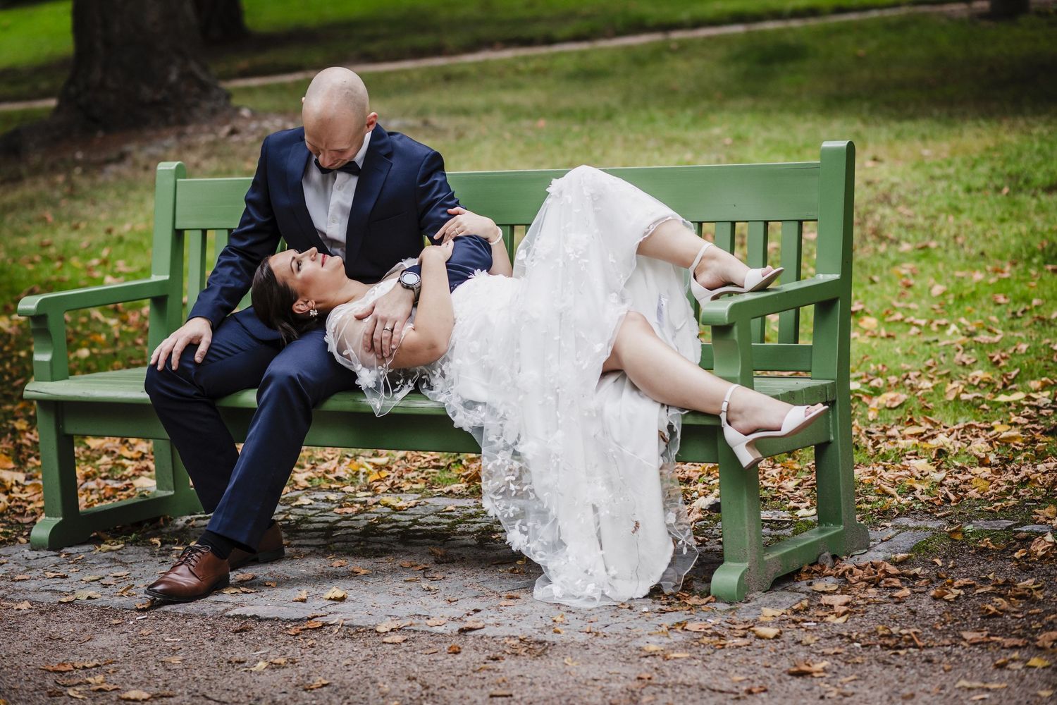 Bride in white lace gown lying across groom's lap on green park bench surrounded by autumn leaves.